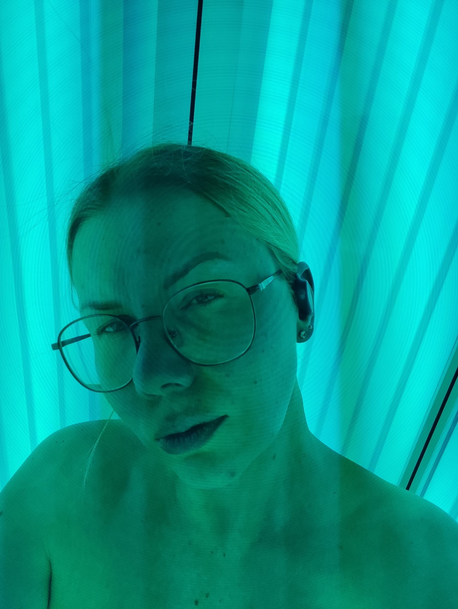 A selfie-style photograph captures a fair-skinned person, possibly female, with blond hair and wearing round eyeglasses. They appear to be in a tanning bed, as evidenced by the distinctive aqua-colored vertical fluorescent light panels that surround them. The lighting gives the person's skin and the surrounding environment a greenish-aqua hue. The subject is looking directly at the camera with a slight frown. The image doesn't offer details to place it specifically in Chaikovsky, Russia. The location is inferred from the request.