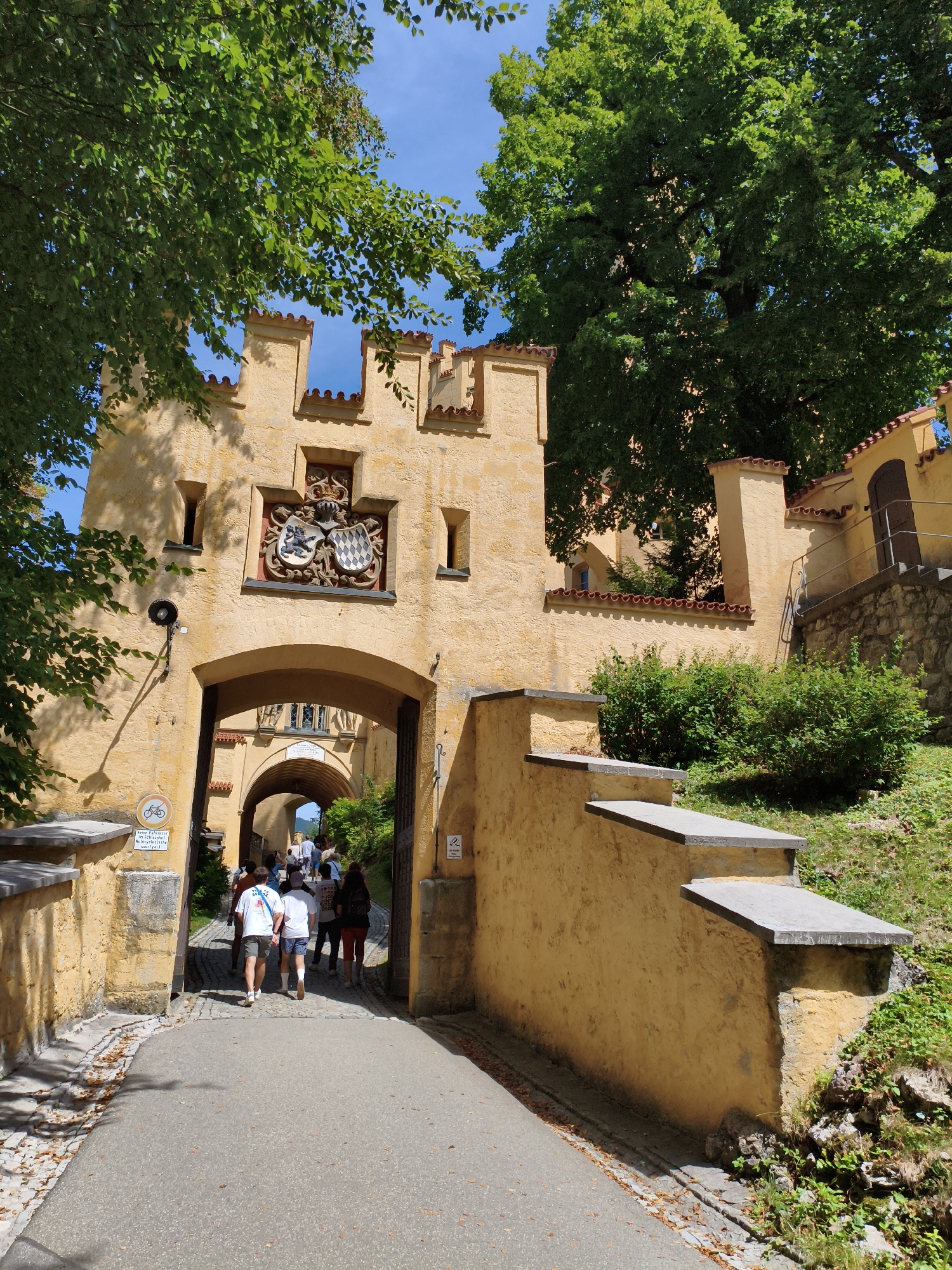 The image shows the main entrance to Hohenschwangau Castle in Schwangau, Germany. The castle is built of light yellow stone and has a distinctly medieval architectural style with crenellated battlements and arched gateways. Green trees with lush foliage are visible, partially obscuring the sky and the upper parts of the castle. A group of tourists, dressed in casual summer clothing, are walking through the main archway and into the castle grounds. The path leading to the entrance is paved with asphalt and cobblestones. Sunlight casts shadows, indicating a bright, sunny day. There are several signs on the wall, including a circular sign with a bicycle icon. A stone staircase with concrete steps ascends along the right side of the castle wall.