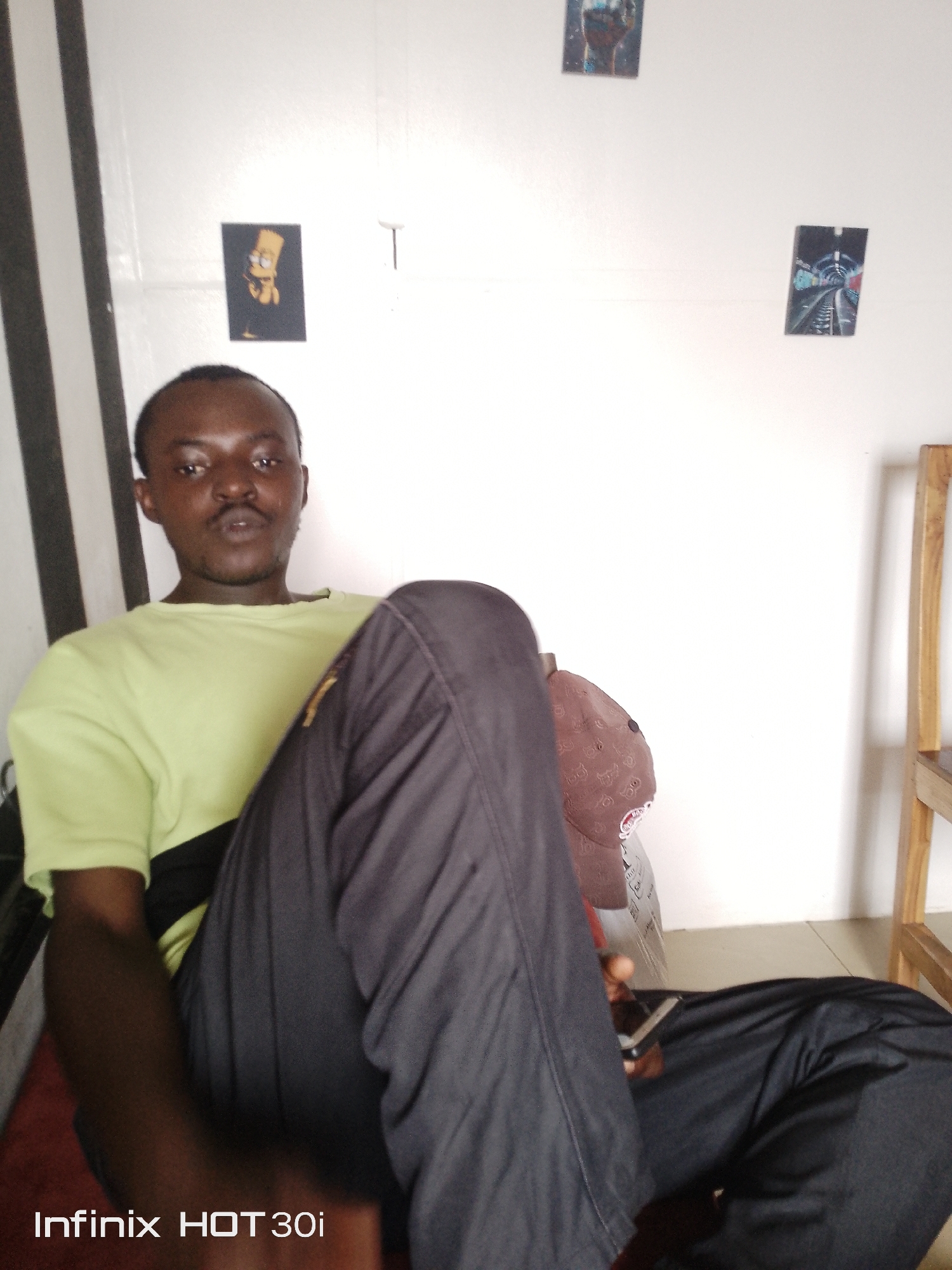 An indoor, eye-level, medium shot shows a dark-skinned male sitting on a reddish-brown surface and leaning back against a white wall. The man is facing the camera with his body angled to the right. He has short, dark hair and a mustache. He is wearing a light green t-shirt with a black band across the chest and dark gray pants. His left leg is bent, and his right leg is extended forward, resting on top of his left leg. He is holding a dark-colored cell phone in his right hand, which is resting on his right leg. A brown baseball cap with a pattern is placed on his bent left leg. The white wall behind him has three small, rectangular pictures affixed to it: one on the left depicting a yellow cartoon character with spiky hair, one in the center with a black background and white lines, and one on the right with a black background and railroad tracks. To the left of the frame, a vertical black and white striped surface is visible. To the right of the frame, a portion of a light-colored wooden shelf unit is visible. The bottom left corner of the image displays the text "Infinix HOT 30i."