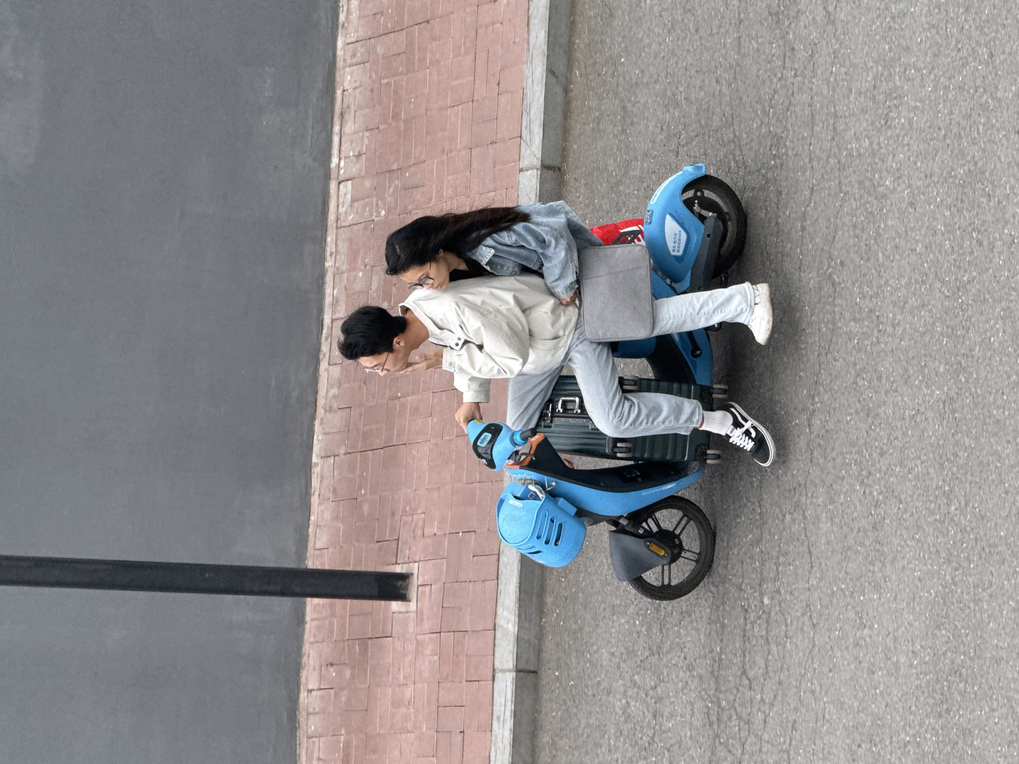 Captured from an elevated perspective, the image shows two individuals riding a light blue electric scooter on an asphalt street bordered by a brick sidewalk and a dark gray wall.
The driver is wearing a white jacket and light-colored pants. The passenger, sitting behind, is wearing a denim jacket. The passenger is holding a gray laptop case.
The environment suggests an urban setting, possibly Zhongtun, China. The sidewalk runs parallel to the street. The scooter is positioned on the right side of the frame. The overhead view focuses on the subjects and their immediate surroundings, revealing details of their clothing, the scooter's design, and the urban infrastructure.