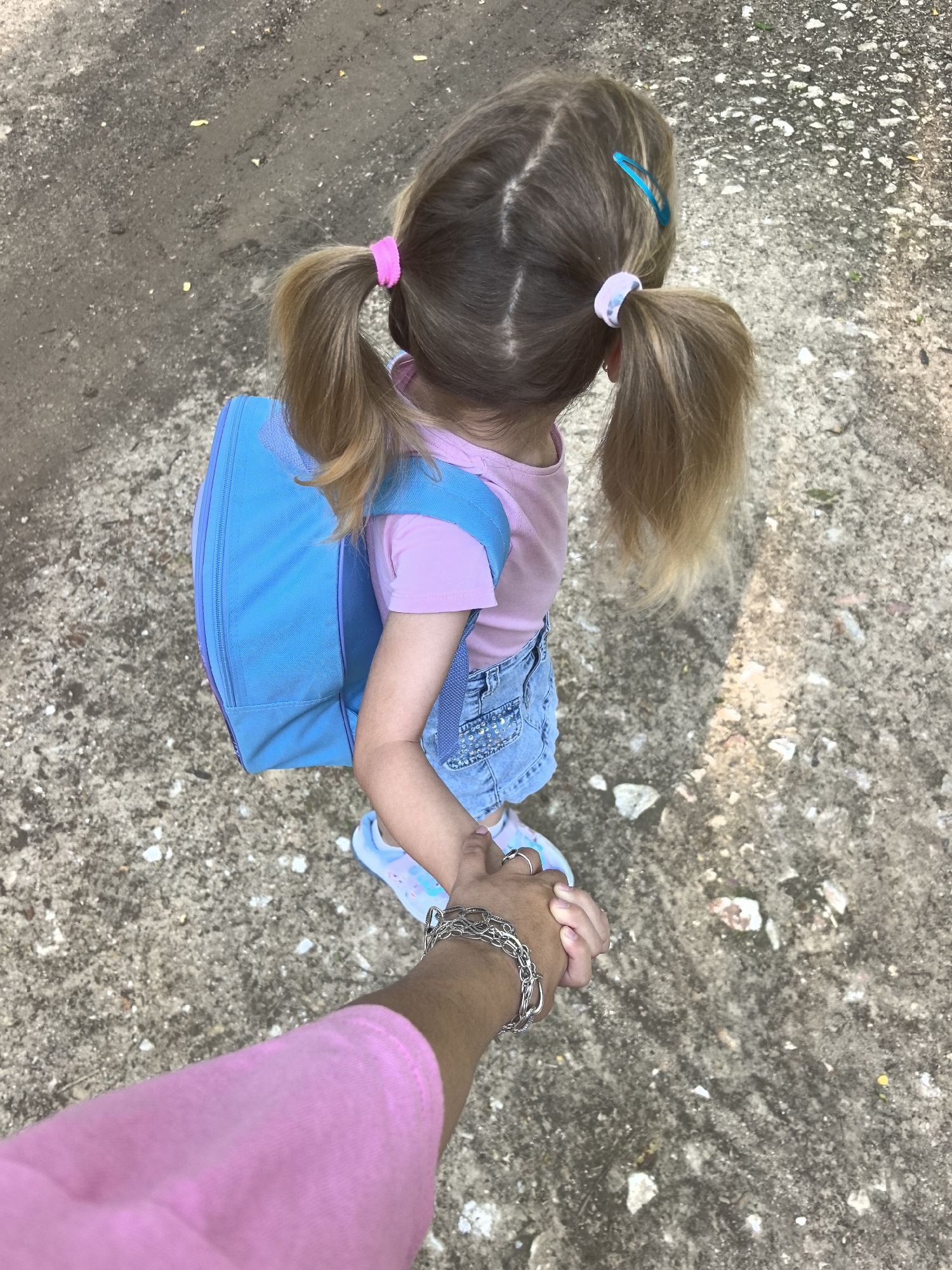 A high-angle shot shows a young girl with blonde hair styled in pigtails, secured with pink and blue accessories. She is wearing a pink t-shirt, denim shorts, and light-colored shoes with a faint pattern. The girl has a blue backpack on. She is holding the hand of an individual wearing a pink shirt and a silver bracelet, likely an adult. The scene takes place on a concrete or paved surface with visible cracks and discoloration. The location is in the city of Plovdiv, Bulgaria.