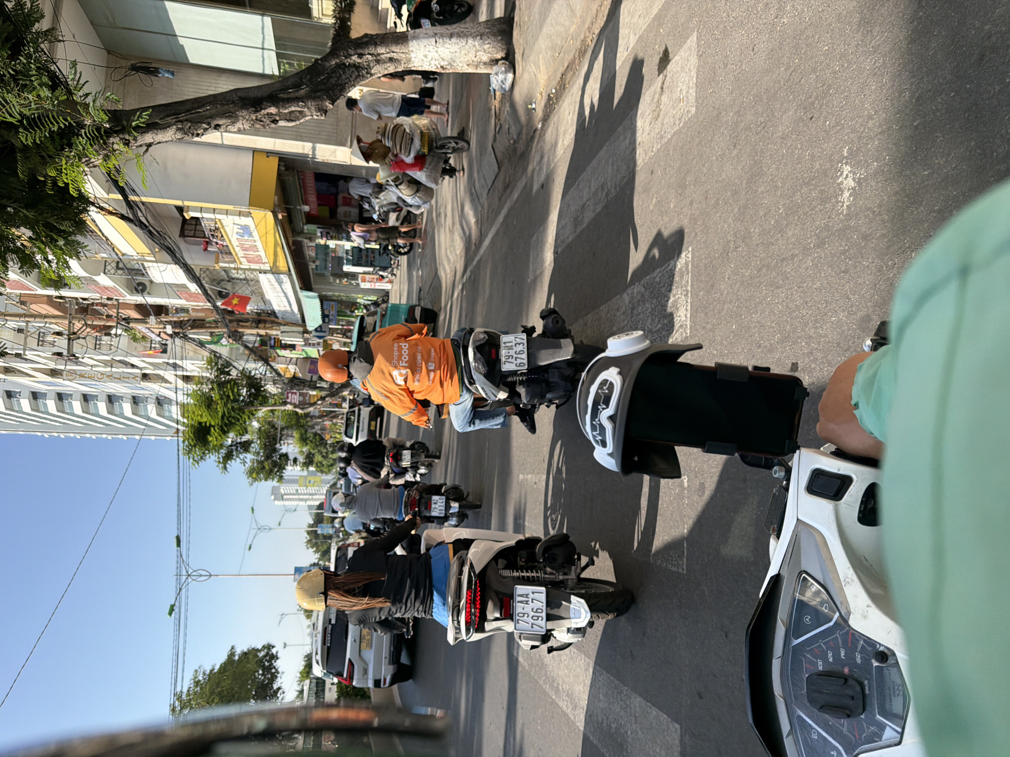 The image captures a street scene in Nha Trang, Vietnam, with traffic and pedestrians. In the foreground, the dashboard and handlebars of a white scooter are visible, with a phone mount attached. A person's arm, wearing a light green shirt, is gripping the handlebar. The scooter is positioned at a pedestrian crossing, indicated by white stripes on the asphalt.
Ahead, several motorbikes are navigating the road. One motorbike, ridden by a person in an orange "Shopee Food" jacket, is in the right lane. Another motorbike, with a rider wearing a yellow helmet and blue shorts, is in the left lane. Further down the road, other motorbikes and a white SUV are visible.
The environment consists of a multi-lane street lined with buildings. On the right side of the street, there are multi-story buildings with shops and balconies. A large tree with visible roots and bark is positioned on the sidewalk. Overhead, power lines and streetlights are strung across the sky. The sky is clear and blue, suggesting a sunny day. Pedestrians are seen on the sidewalk and riding on motorbikes. One individual on a motorbike is carrying a large load of goods. A Vietnamese flag is displayed on one of the buildings. The overall impression is a busy urban street with typical traffic and commercial activity in Vietnam.