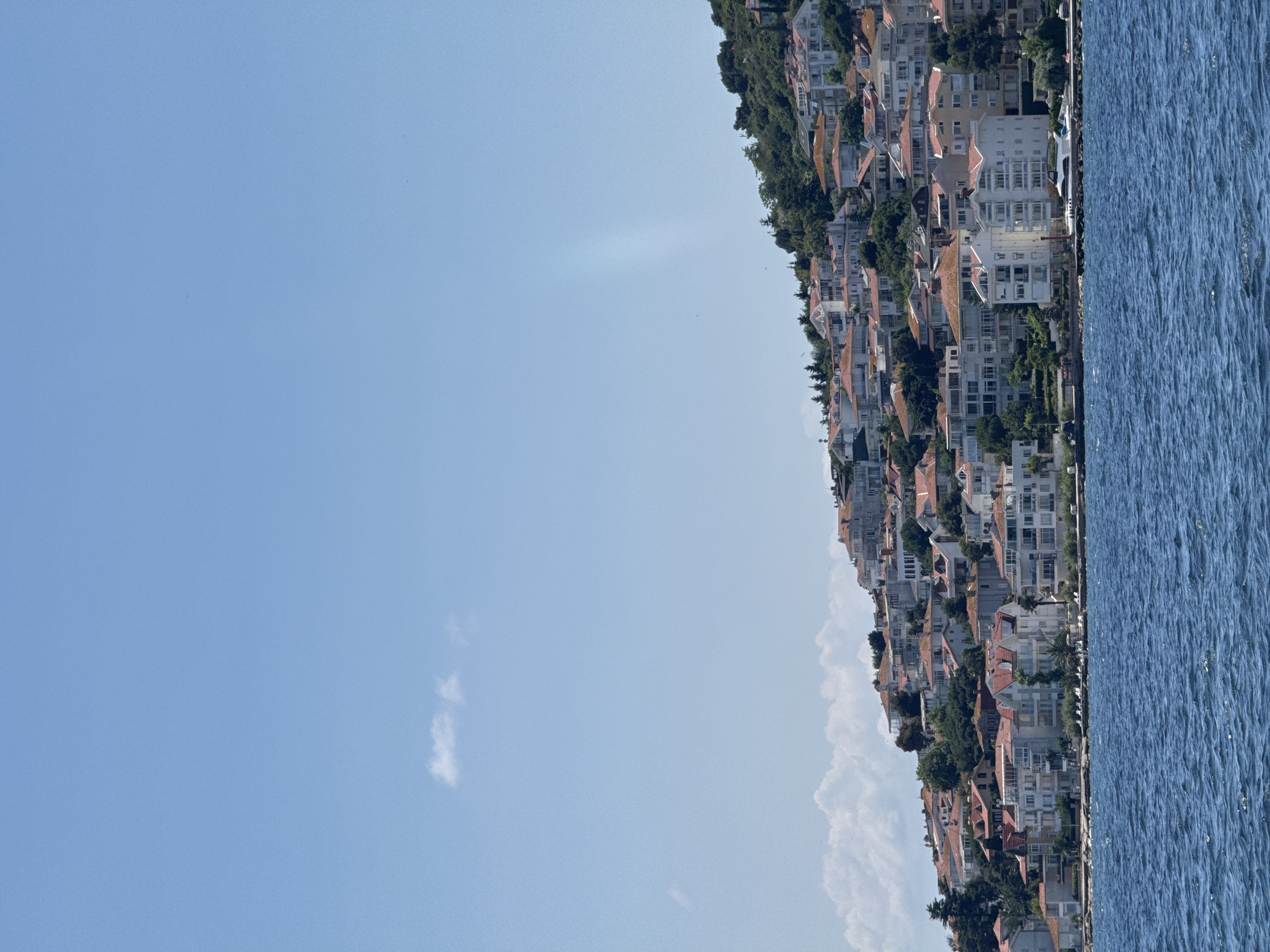 This is a photograph of the city of Prince Islands, Turkey, taken from the water. The foreground is dominated by the choppy blue water of the sea, with whitecaps visible. In the background, a hillside is covered with a dense arrangement of buildings, characteristic of island architecture. Many of the buildings feature red-tiled roofs and white or light-colored facades. Some of the structures are larger apartment buildings, while others appear to be more traditional homes. Lush green trees are interspersed among the buildings, indicating a verdant landscape. The sky above is a clear, pale blue with a few wispy white clouds. A single, long white yacht is docked along the shoreline in front of some of the buildings. There are no visible people in the image.