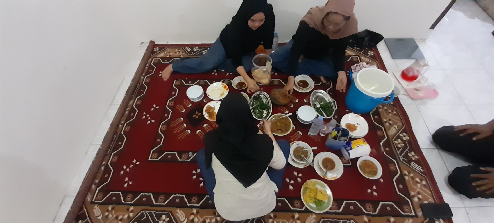 In the city of Gondang, Indonesia, four individuals are captured in a high-angle shot, seated on a red and brown patterned rug, engaged in a meal. The scene appears to be an indoor setting, possibly a dining room, with white walls and tiled flooring visible.
Three women are the primary subjects, all wearing hijabs. The woman on the far left, wearing a black hijab and blue jeans, is actively reaching for food. Next to her, a woman in a light brown hijab and dark top also participates in the meal. In the foreground, another woman, wearing a black hijab and a white top, is seated and looking down at the food. The fourth individual, on the far right, is partially visible with dark clothing, also seated.
The rug is laden with various dishes: several white plates with food, small bowls containing sauces or curries, and a large metal platter with green vegetables. A large blue insulated container, likely for drinks, is positioned on the rug. Other objects present include plastic containers, water bottles, and a large, dark, curved object that could be a serving dish or a piece of fruit.
The overall atmosphere suggests a casual, communal dining experience. The lighting is neutral, with no harsh shadows, indicating ambient indoor lighting. The composition emphasizes the shared meal and the interactions between the individuals.
