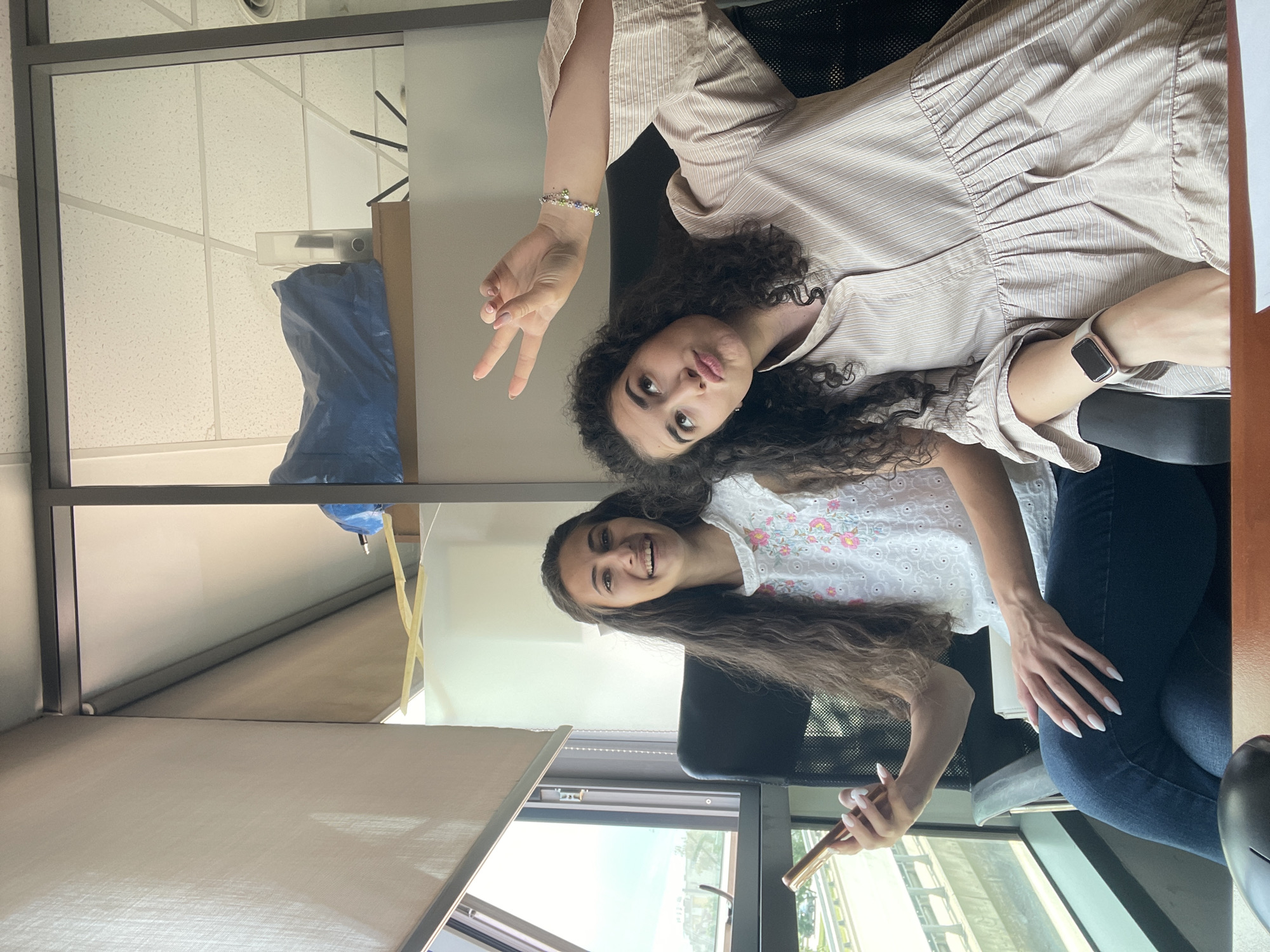Two young women are seated in an office environment. The woman on the left has long dark curly hair and is wearing a white shirt with floral embroidery and dark blue jeans. She is holding a gold-colored smartphone in her left hand. The woman on the right has dark curly hair and is wearing a light brown pinstripe dress. She is making a peace sign with her right hand. The office appears to be a modern workspace with glass partitions. Behind the women, shelves are visible with some items, including a blue tarp and files. Through a large window on the left, a glimpse of the city can be seen, suggesting an urban setting. The presence of blinds on the window indicates control over natural light. The overall scene suggests a casual moment captured within a professional or educational setting. This could be an office building located in the city of Sofia, Bulgaria, given the general architectural style and the possibility of an urban landscape outside the window.