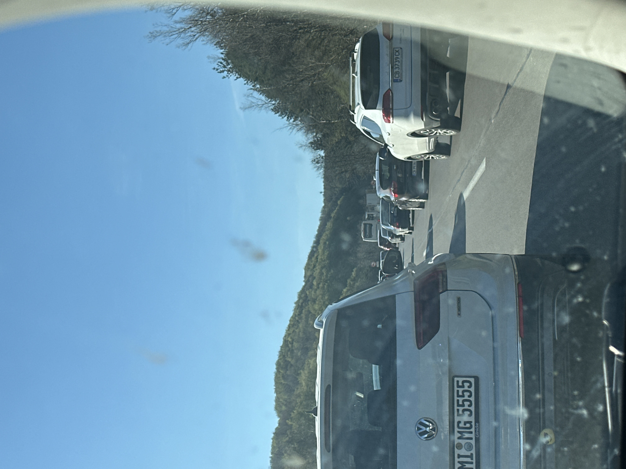 The image shows a line of cars in traffic, likely on a road leading to or from Dolna Vasilitsa, Bulgaria. The car in the foreground is a light-colored Volkswagen with the license plate "MI MG 5555." Behind it, several other vehicles are visible, including a white car with the license plate "CB 3739 CK." The environment includes a clear blue sky and a hillside covered with trees. The perspective suggests the photo was taken from inside a vehicle.