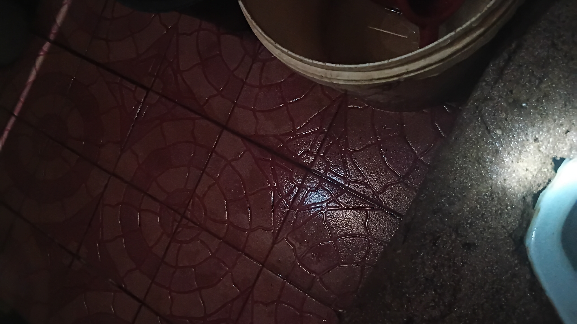 The image depicts a wet, tiled floor with a circular, patterned design in a deep red hue. The tiles are arranged in a grid, and water is visible pooling on the surface, reflecting the dim lighting. In the lower right portion of the frame, the rim of a light-colored bucket is visible, with some red objects inside. To the lower left, a white object, possibly a toilet bowl, is partially visible, illuminated by a bright light source. The overall scene appears to be an indoor environment, possibly a bathroom or utility area, with evidence of recent spillage or cleaning. The location of Prabumulih, Indonesia is not discernible from the visual information provided in the image.