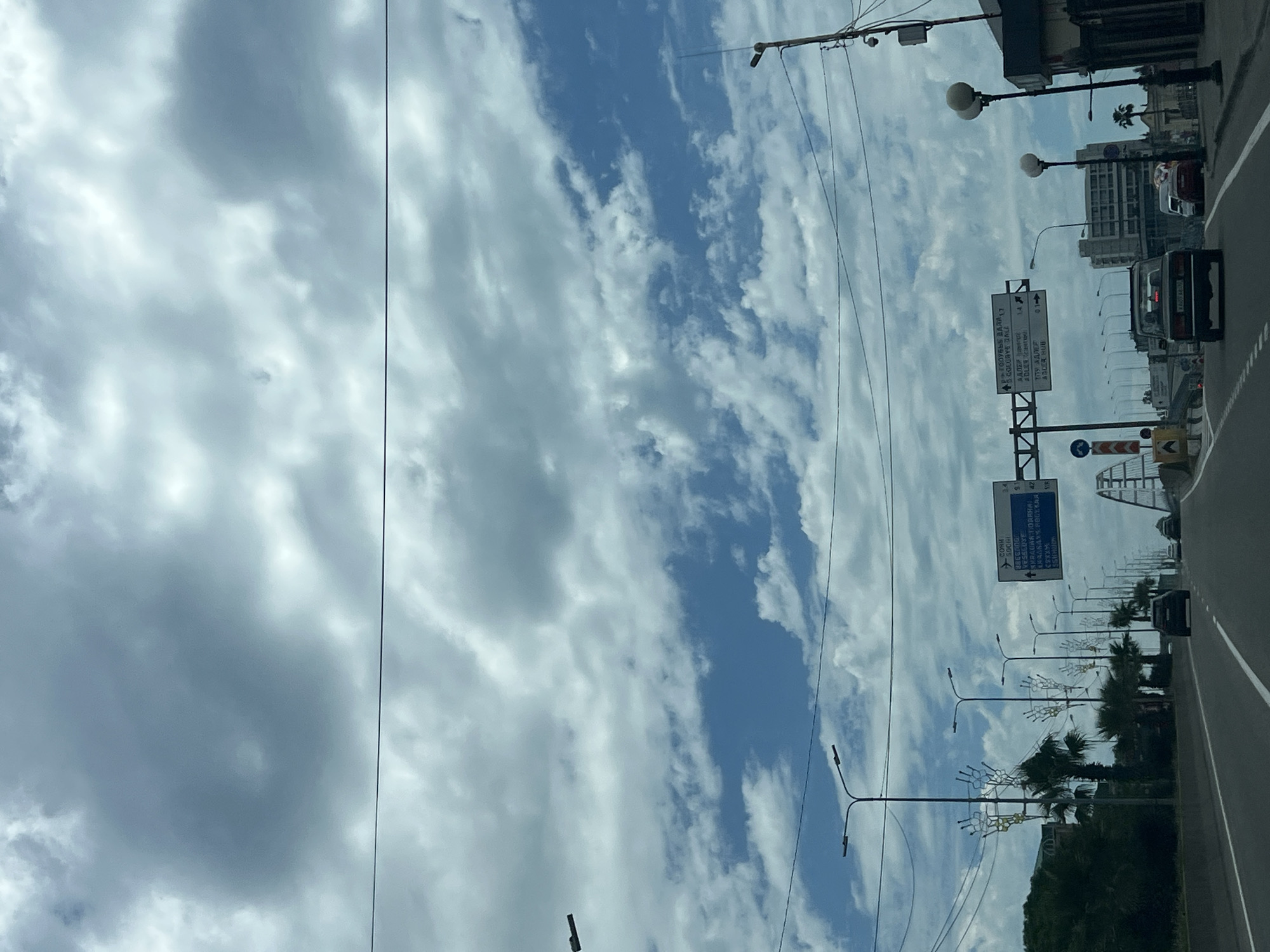 The image shows a view from a vehicle traveling on a highway with a cloudy sky. There are multiple streetlights and palm trees lining the road. Road signs are visible, with some indicating directions to "Сочи" (Sochi) and "Адлер" (Adler). Several cars are on the highway, and buildings can be seen in the background. The overall environment appears to be urban or suburban.