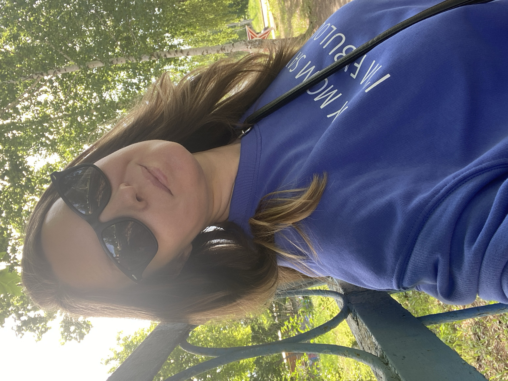 A woman in sunglasses and a blue sweatshirt with white lettering is leaning on a blue metal railing. Trees are visible in the background, suggesting a park setting. A red and white object is also visible in the background. The scene is in Noviy, Russia.