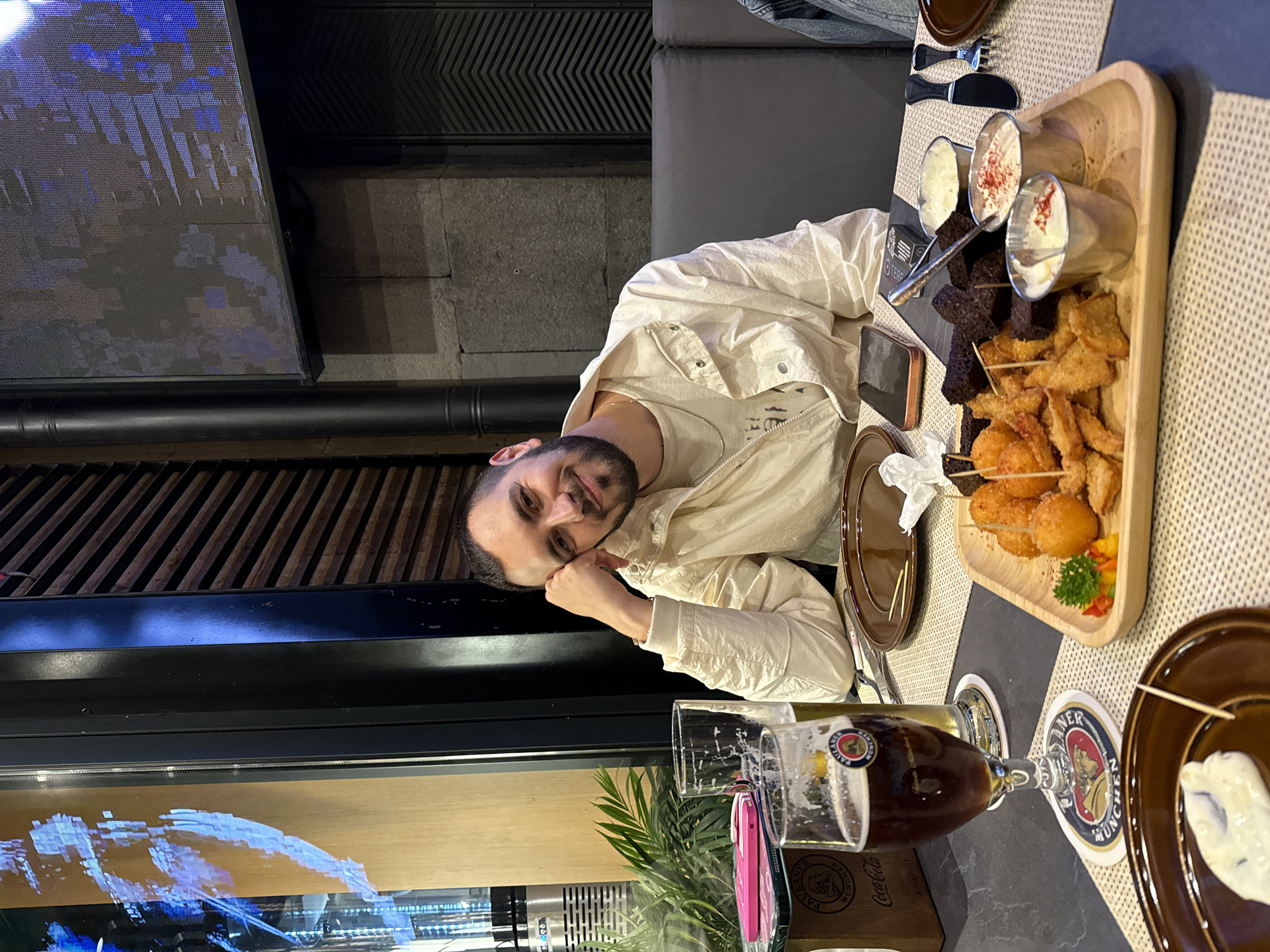 The image depicts a man seated at a table in what appears to be a restaurant or bar. The table is covered with a textured beige tablecloth and has various items on it including a wooden tray with a selection of fried foods and dipping sauces, a plate with a white dip, cutlery, and two Paulaner beer glasses, one partially filled with dark beer. A Coca-Cola branded container is also visible. The man is leaning on his hand and looking at the camera. Behind him, there's a dark-colored wall with a decorative panel and a window or glass partition displaying a screen with blue and white patterns. A potted plant is seen in the lower part of the frame. The style of the establishment and the presence of Paulaner beer suggest that this is a restaurant. I am unable to confirm that this is Yerevan, Armenia.