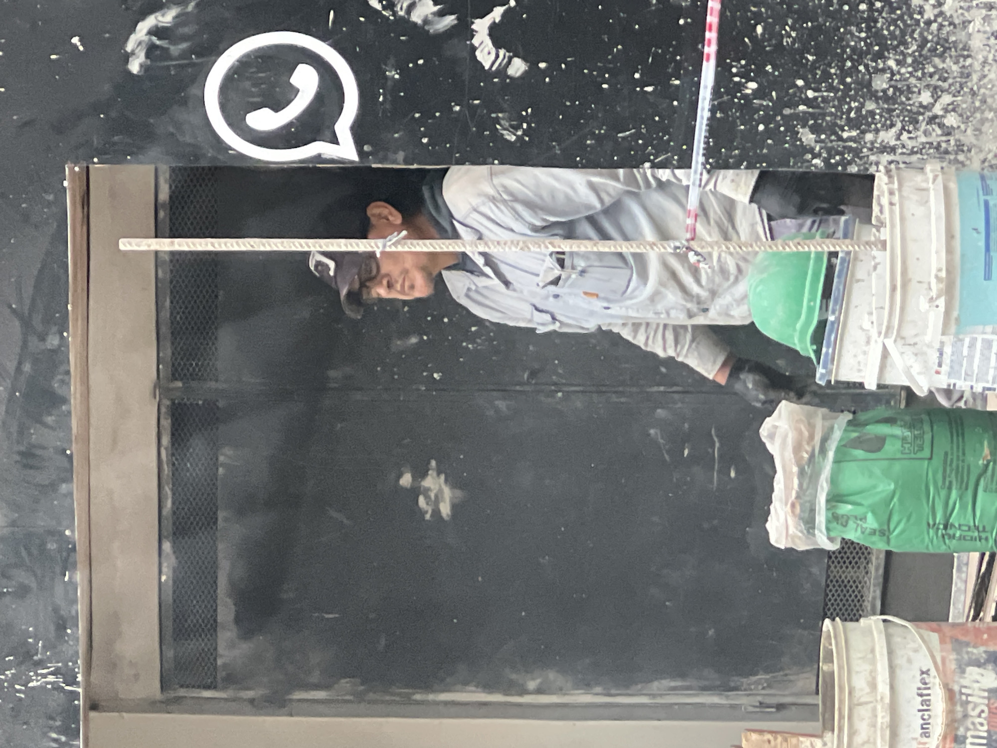 The image shows a construction worker with a green helmet and work clothes standing in a doorway below a black surface with a white WhatsApp logo. He is working with construction materials. He is holding a metal trowel and is in front of several buckets. The location is Buenos Aires, Argentina.
