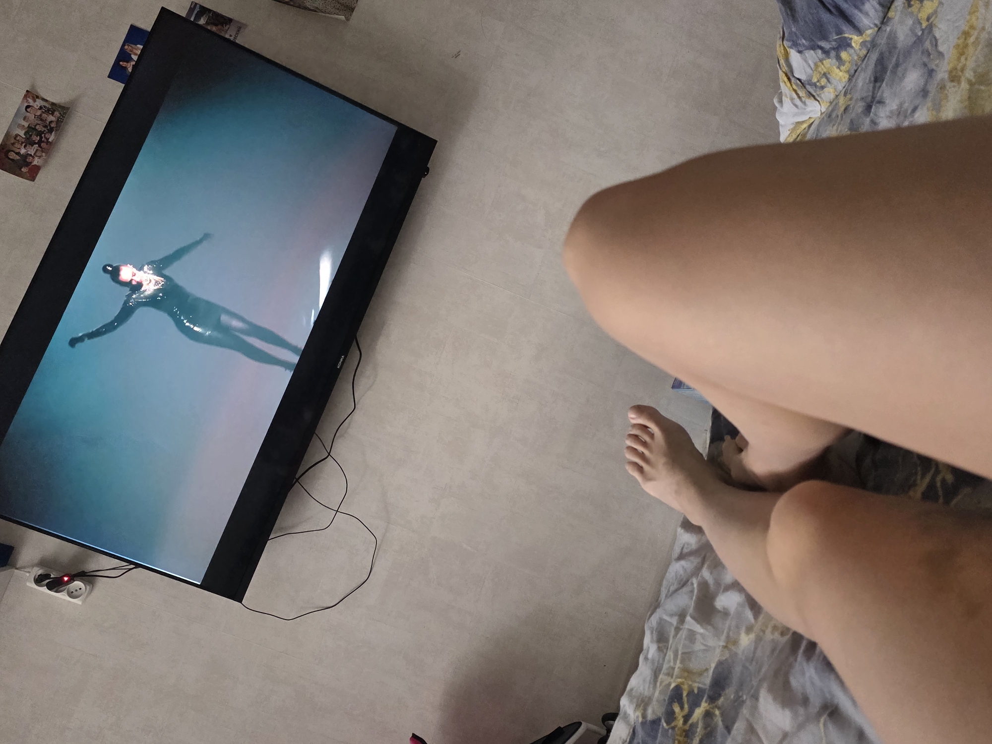 The image shows a person lying on a bed with a television in the background. The television is displaying an image of a person in a wetsuit underwater. The person on the bed has their legs visible, and a foot is partially visible. The bed has a patterned grey and yellow duvet. The floor is a light grey tile. There is a power strip visible near the television. There are also various small cards or pictures near the television. There is no information to determine the location is Chaikovsky, Russia.