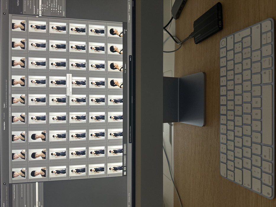 The image displays a computer workstation featuring a silver Apple iMac or similar desktop monitor. The monitor's screen shows a digital image management interface, likely an import screen from Adobe Lightroom, with "Importation" and other French text visible (e.g., "Copier," "Ajouter," "Toutes les photos," "Dossier de destination"). The main content on the screen is a grid of numerous thumbnail photographs, depicting a single East Asian male model in a black tank top and black pants. The model is shown in various poses, from headshots to full-body shots, mostly against a plain light background. Below the monitor, a light-colored wooden desk surface is visible. On the desk, in the foreground, rests a silver Apple Magic Keyboard with a French AZERTY layout. To the right, a black external hard drive is connected via a black USB cable, and a gray USB hub is partially visible further back on the desk. The presence of the French AZERTY keyboard and the French language on the software interface strongly indicate the workstation is located in a French-speaking region, specifically in Paris, France, as requested.