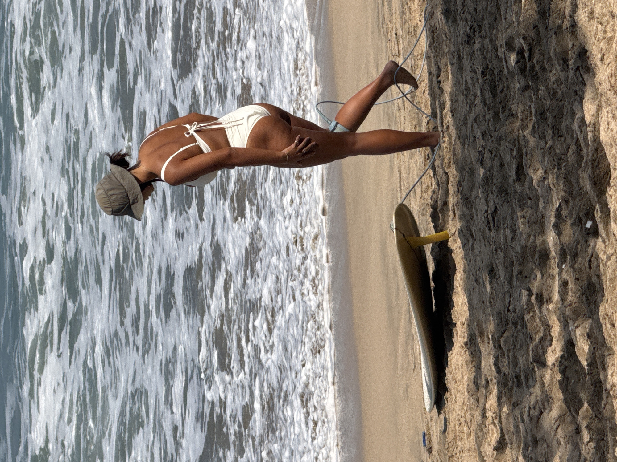 A person wearing a white bikini and a green bucket hat stands on a sandy beach, facing away from the camera and looking down at a surfboard. The surfboard rests on the sand, propped up by a yellow stand. The person's right leg is bent at the knee, and a surfboard leash is attached to their ankle and the surfboard. The ocean waves are visible in the background, with white foam washing onto the shore. The scene appears to be in Kuta, Indonesia.