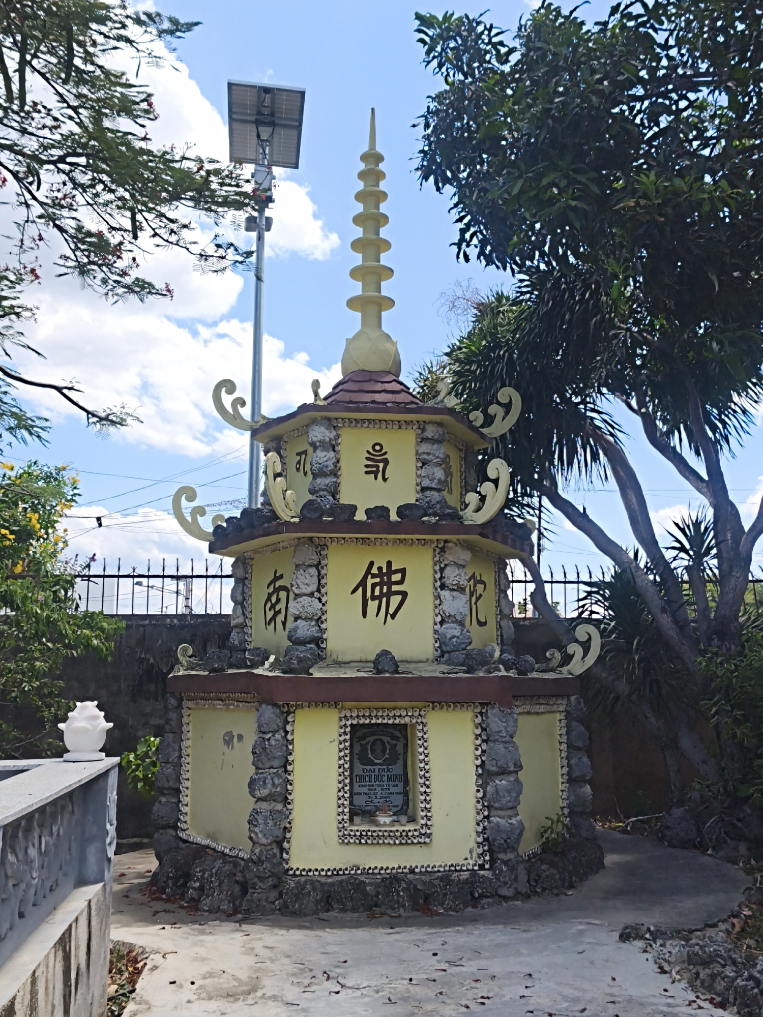 The image shows a multi-tiered, yellow pagoda structure, possibly a shrine or memorial, adorned with decorative stone and seashell embellishments. The pagoda is topped with a finial and features ornate scrollwork on its eaves. Chinese characters are visible on its upper sections. In front of the pagoda, a small plaque with Vietnamese text is set within a framed niche. The structure is situated outdoors, with a fence and trees in the background. To the left, a grey concrete wall with carved details and a white decorative object are present. A solar-powered lamp post stands behind the pagoda. The ground is a mixture of paved areas and dirt, with fallen leaves scattered around. The environment suggests a warm, possibly tropical climate with clear skies and some clouds. The presence of Vietnamese text on the plaque strongly indicates the location as Vietnam. Given the context of pagodas and memorials, and the specific inscriptions, this likely depicts a Buddhist site in Cam Ranh, Vietnam.