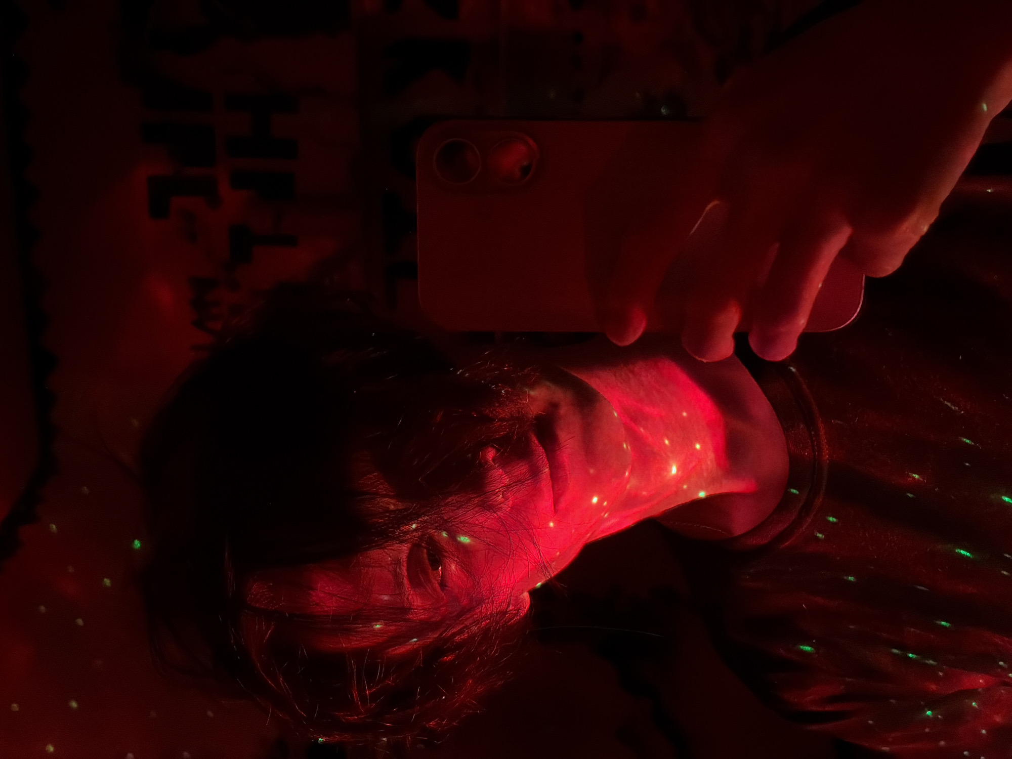 A person, whose face is partially obscured by shadows and red light, holds up a smartphone in their right hand, taking a selfie. The lighting is predominantly red, with small green specks scattered across the image, suggesting a festive or atmospheric setting, possibly a party or a room decorated with lights. The person appears to be wearing a dark-colored t-shirt. In the background, dimly visible text or graphics are present on what appears to be a wall, but they are indistinct. The overall mood is intimate and somewhat mysterious due to the low-light conditions and the dramatic lighting. The location is Unknown, Unknown.