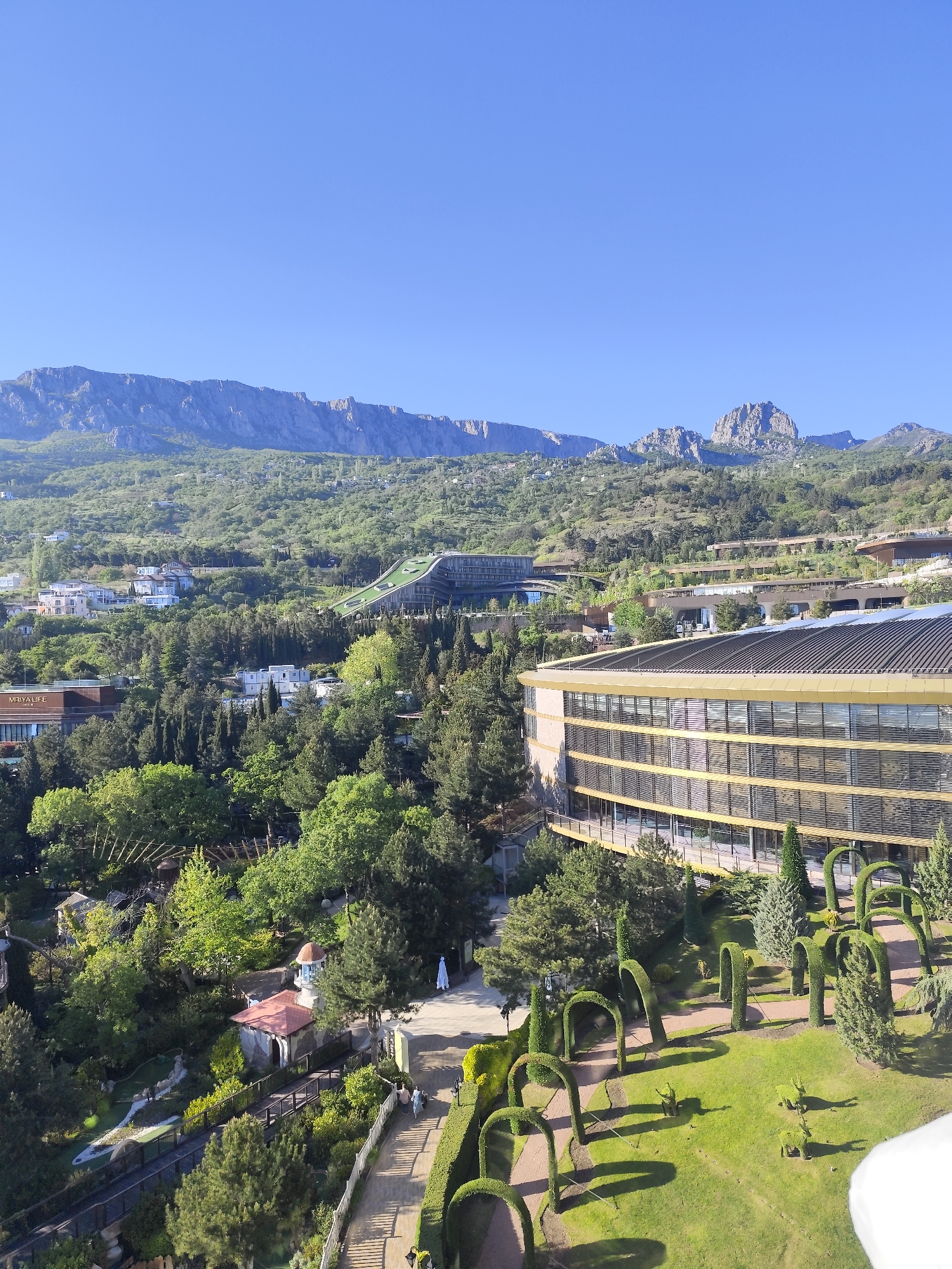 The image shows a landscape view in Симеизский поселковый совет, Ukraine. In the background are mountains under a clear blue sky. The midground features a green, hilly landscape with trees, buildings, and what appears to be a hotel or resort complex with a curved structure. In the foreground, there is a manicured garden with arch-shaped shrubs and green lawns. A pathway winds through the garden, and there are a few people visible walking along it. Overall, the scene depicts a blend of natural mountain scenery and designed landscape elements within the Ukrainian town.