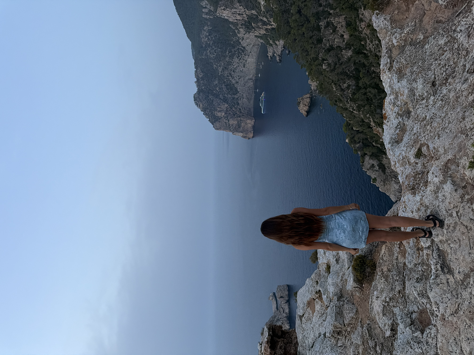 A woman stands on a rocky cliff overlooking a calm, deep blue sea. She is viewed from behind, facing away from the camera, with her long, dark brown hair flowing down her back. She is wearing a short, light blue denim-like dress and dark sandals. The cliff edge is rough and textured, with sparse vegetation. In the distance, a large, multi-story yacht is anchored in a cove, accompanied by several smaller boats. The surrounding landscape features steep, rocky cliffs covered in dark green pine trees. The sky is overcast and a muted blue-grey, indicating dusk or a cloudy day. The scene suggests a coastal view, likely in a Mediterranean location, given the natural formations and the style of the yacht. The location is near Sant Antoni de Portmany, Spain, where similar dramatic cliffside views of the Mediterranean Sea are common.
