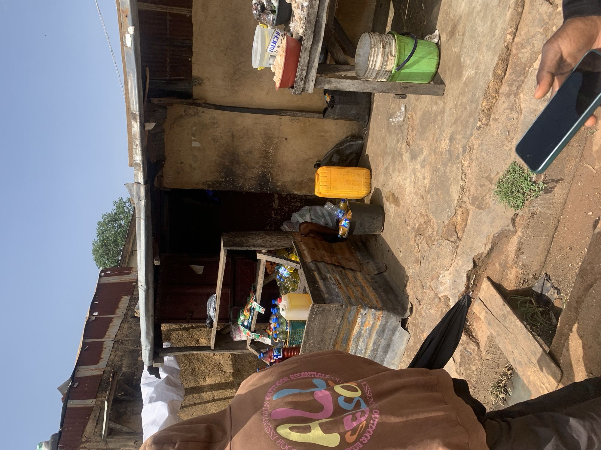 The image depicts a street scene in Gura Top, Nigeria. There is a small, informal shop or stall selling various items, including bottles and containers. The stall appears to be constructed of corrugated metal and wood. A person is holding a smartphone. The environment is somewhat cluttered and uneven, suggesting a busy, possibly impoverished area. The lighting indicates it is daytime.