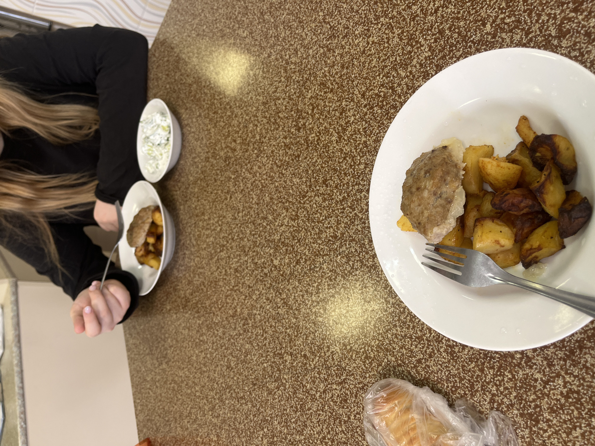 A high-angle indoor shot shows a person sitting at a brown speckled tabletop, with food arranged in two white bowls and on a white plate. The person on the left is partially visible, wearing a black top and having long blonde hair. Their hands are holding a fork, and they are eating from a bowl that contains fried potatoes and what appears to be a meat patty. Next to the bowl is another white bowl filled with a white salad. On the right of the frame is a white plate with a meat patty and fried potatoes. A fork is placed on top of the potatoes. In the bottom right corner, a bread roll is wrapped in plastic. The location appears to be a cafeteria or simple restaurant. Based on the food served, and the style of the cafeteria, it is possible this photograph was taken in a cafeteria in Saint Petersburg, Russia.