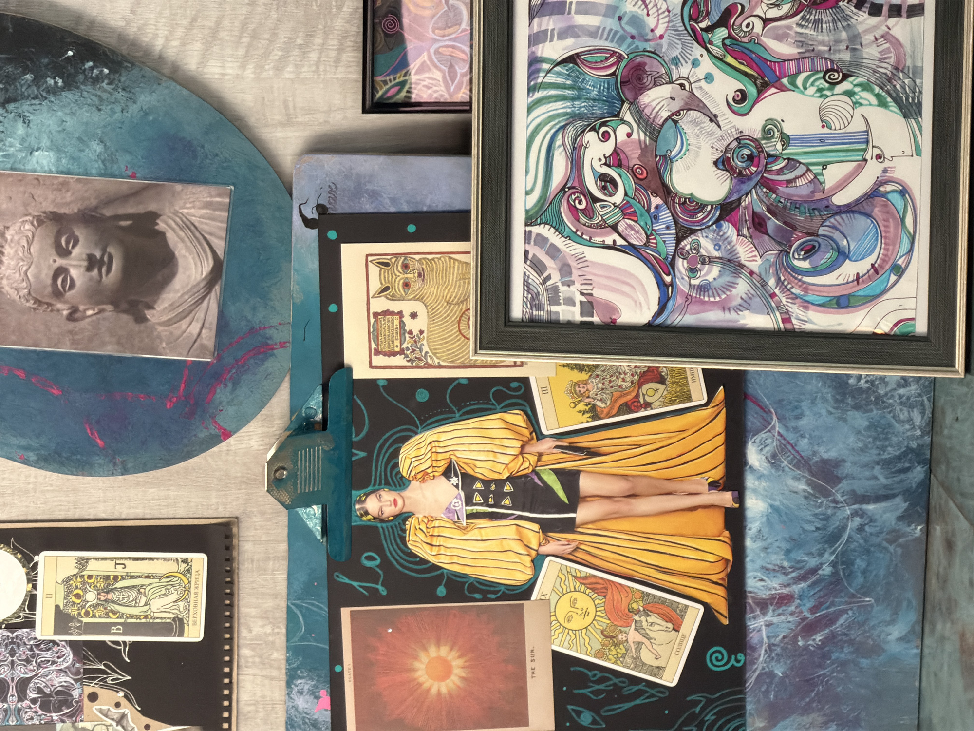 The image shows a collection of art pieces and ephemera arranged on a surface with a light gray wood grain pattern.
On the left, there is a circular object with a painted blue and indigo surface, with a small square photo of a Buddha statue adhered to it. To the right of this is a stack of layered items. At the bottom of the stack, there is a blue-toned notebook or art pad, held together with a blue clip. The page is adorned with a collage including a photograph of a woman in a yellow dress, tarot cards ("The Sun" and another), and an image of the sun. Above this, another layer reveals an illustrated drawing of a cat.
The uppermost right portion of the image features a framed abstract art piece with intricate line work in purple, blue, and white, on what looks to be a painting on canvas.
A small, black notebook with spirals is in the lower left corner, and a tarot card is on top of it.