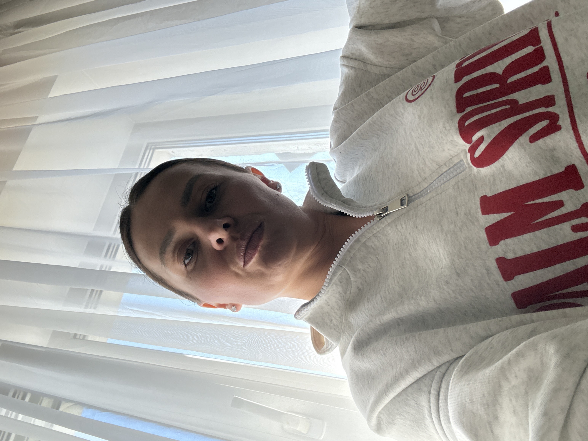 A selfie taken indoors. A woman with short, dark hair and light skin looks directly at the camera with a neutral expression. She wears a gray zip-up sweatshirt with red lettering. The background is a window with white sheer curtains. The location is unknown and cannot be identified as Chaikovsky, Russia from the provided image.