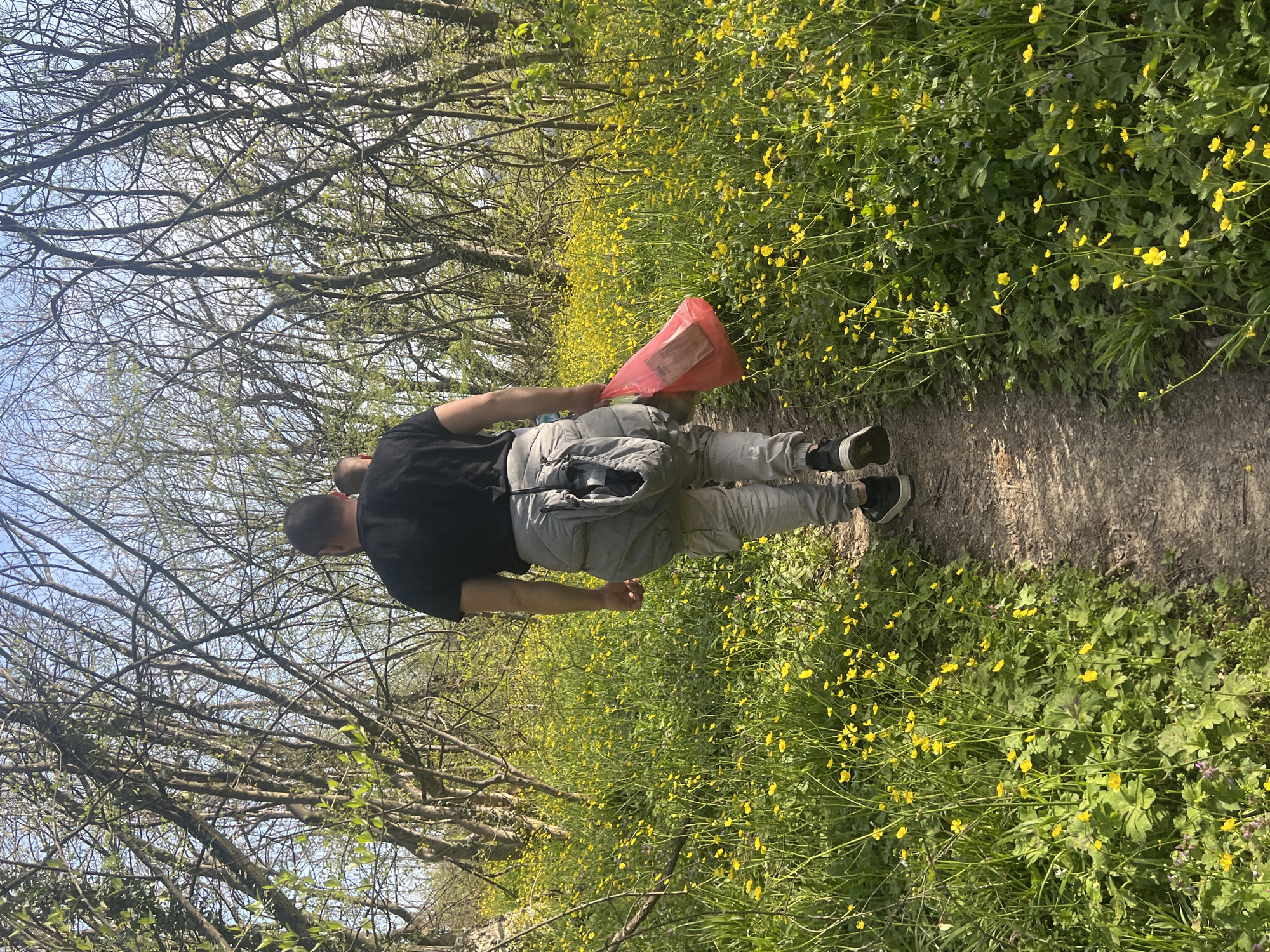 A man walks along a dirt path in Samovodene, Bulgaria. He wears a black t-shirt, light gray pants, and dark-colored shoes. A gray jacket is tied around his waist. He carries a red plastic bag in his right hand. The path is bordered by dense vegetation, including flowering plants with yellow blossoms. Bare trees line the area beyond the vegetation on both sides of the path. The sky is visible through the branches.