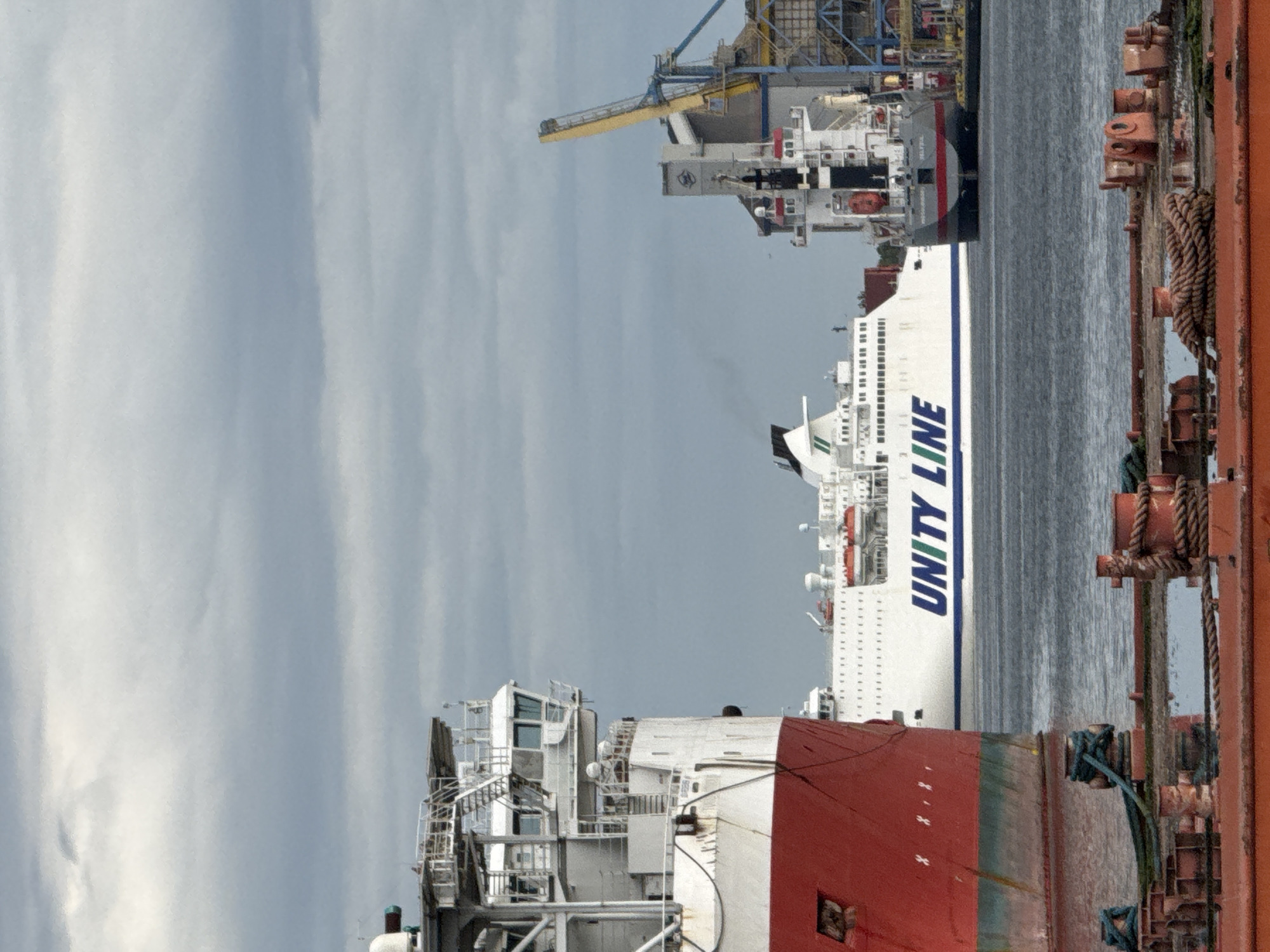 The image depicts a harbor scene in Gdańsk, Poland, featuring several ships and port structures under a cloudy sky. In the foreground, a large red and white ship is docked, with its hull painted red and its upper structure white. Mooring ropes are visible on bollards in the foreground. In the mid-ground, a white ferry with "UNITY LINE" written in blue on its side is sailing. Behind the ferry, another ship, a cargo vessel, is docked. To the right, a tall yellow crane rises above industrial buildings, indicating a working port environment. The water in the harbor is calm and reflects the muted colors of the sky and vessels. There are no people clearly visible in the image. The overall atmosphere is one of industrial activity and maritime transport.
