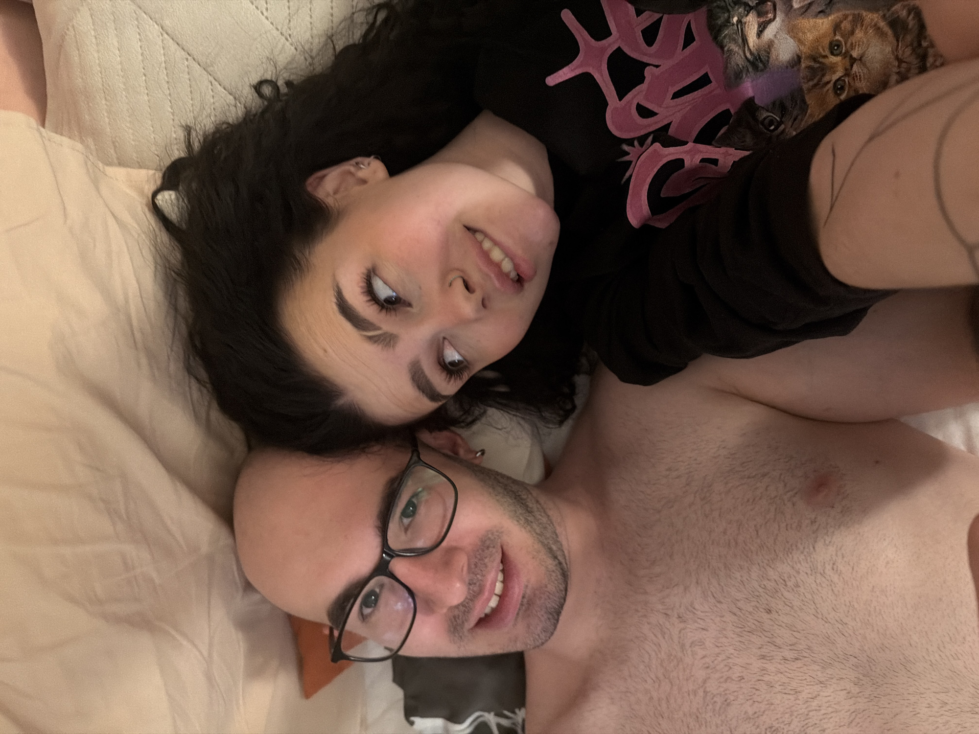 A photograph depicts a couple lying on a bed with light-colored bedding. The woman has dark, curly hair and is wearing a black long-sleeved shirt with a pink graphic design of kittens. The man is bald, has a beard, and is wearing glasses; he is shirtless. Both are smiling and looking towards the camera. The location appears to be indoors, possibly a residential bedroom. There are no apparent indicators to confirm a location in Kazan, Russia.