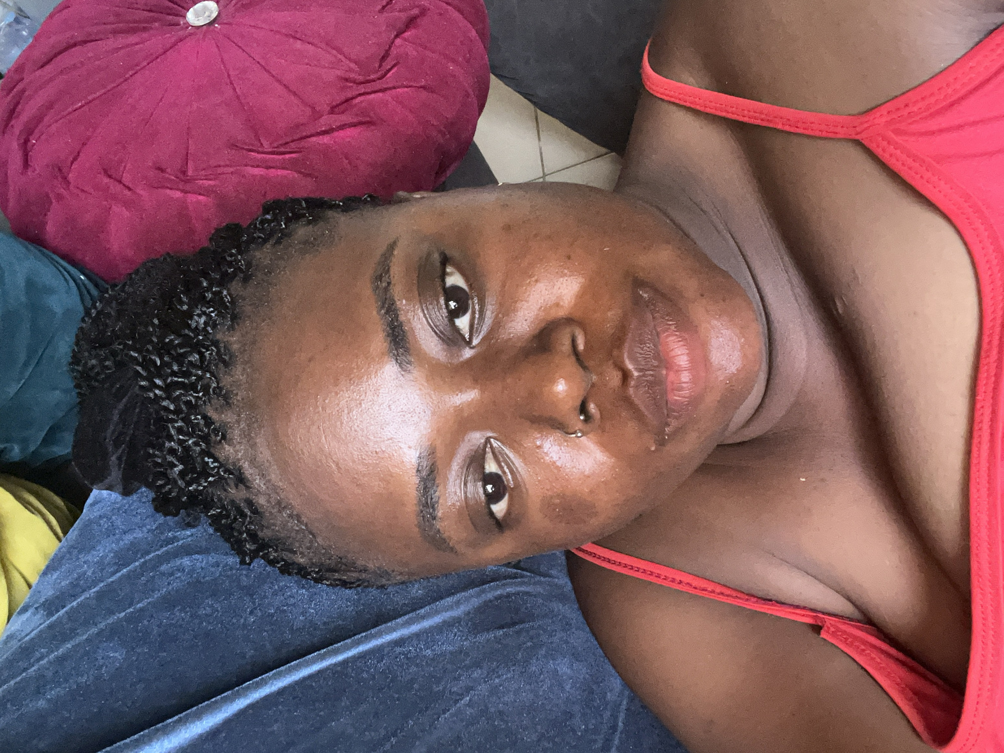 This is a close-up, overhead selfie of a Black woman in Abuja, Nigeria, taken indoors. The woman has dark skin, short, braided hair pulled up, and is wearing a red tank top. She is lying on a surface that appears to be a couch or bed, with a blue fabric visible to her left and a large, round, magenta pillow to her right. The lighting suggests it is daytime, and the image is slightly overexposed, with some shine on her skin. The background is out of focus.