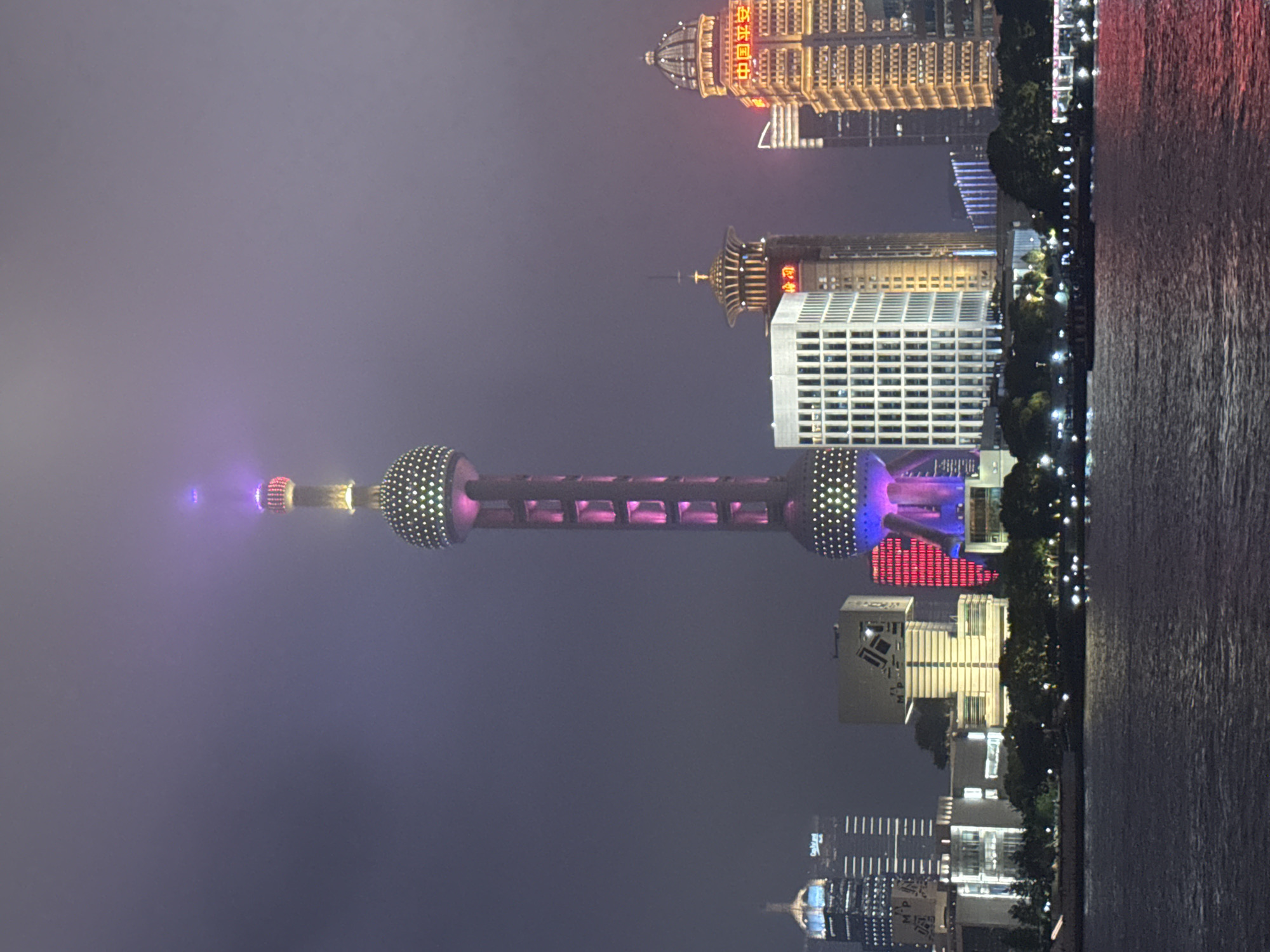 Captured at night, the image presents a skyline view of the Huangpu District in China. At the forefront, the Oriental Pearl Tower stands out, adorned with illuminated spheres connected by a central structure. A river runs along the right edge of the frame, its surface subtly reflecting the cityscape lights. Buildings of various heights and designs form the cityscape, some with distinct architectural details and lighting schemes. The sky is shrouded in a hazy, dark ambiance, casting a subdued glow over the urban environment.