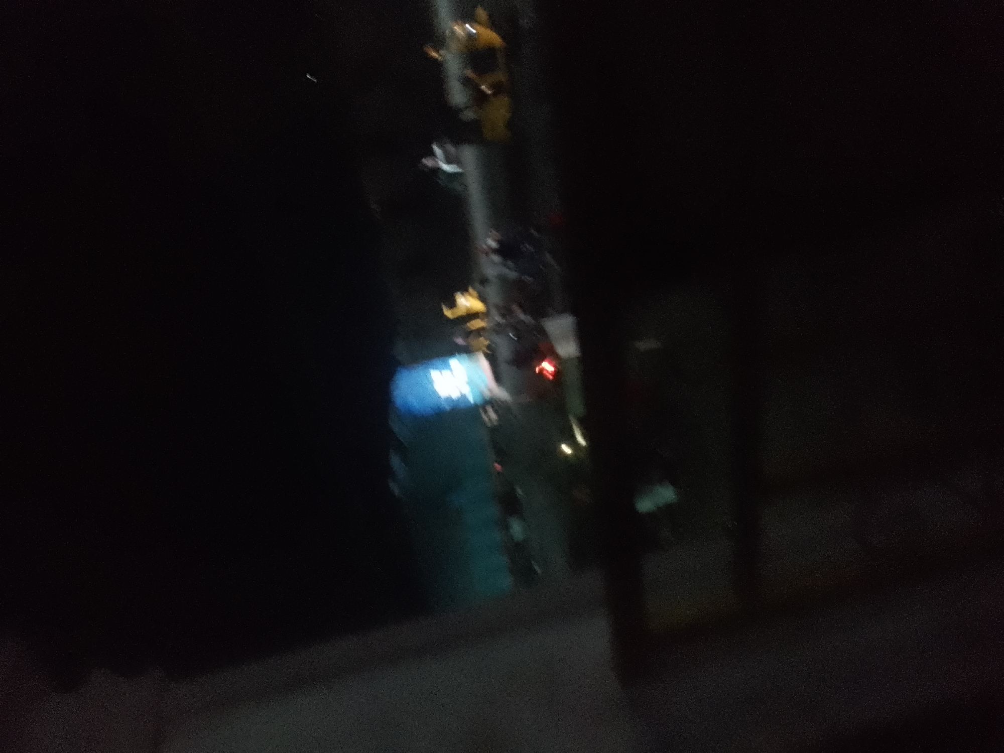 This is a dark, blurry, and out-of-focus photograph taken at night, likely through a window, showing a street scene in Jalingo, Nigeria. The primary subject is a traffic light, with its yellow and red lights visible. There are also blurred figures of people in the foreground, appearing to be walking or standing. The background is dark, with some distant lights suggesting it's an urban environment. The overall image quality is poor due to low light conditions and motion blur.