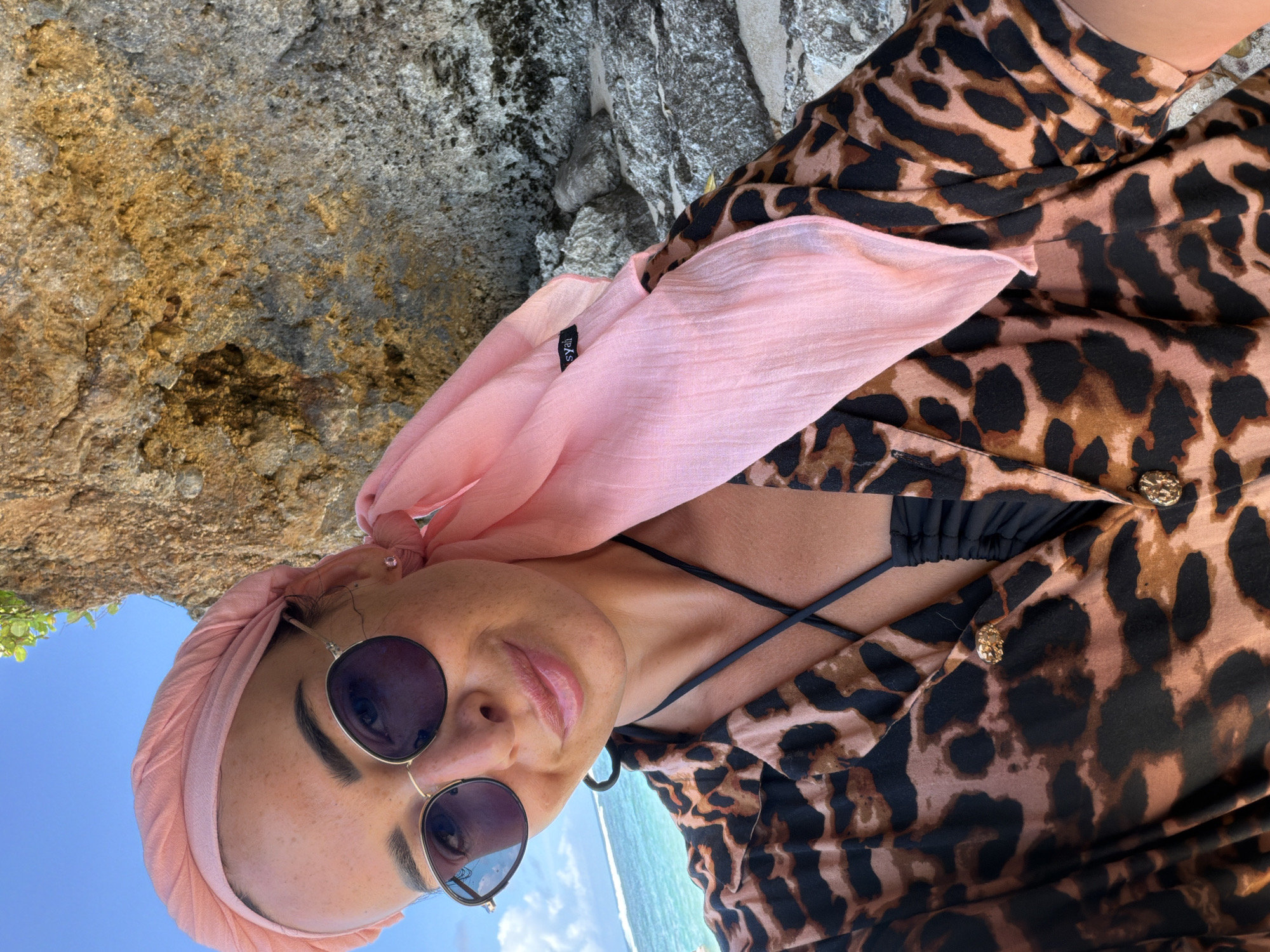 A woman is seen wearing a pink headscarf and sunglasses, and a brown-and-black leopard print dress. Her dress has gold buttons, and she is wearing a black bikini underneath. The ocean is visible in the background, along with some trees and blue sky. She appears to be standing near a large rock. The location is Kutuh, Indonesia.