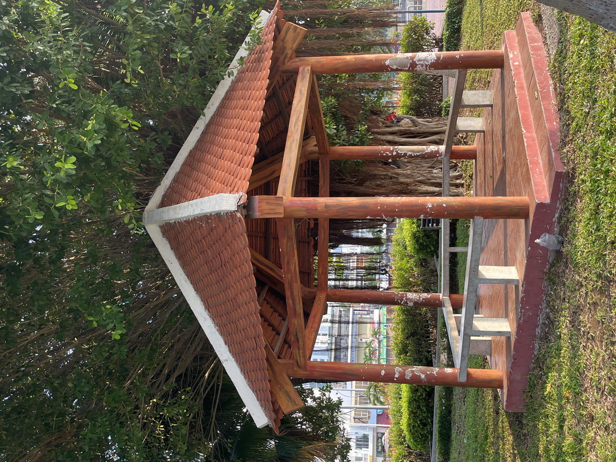 A photograph depicts a red-roofed, wooden gazebo situated in a park, oriented so that the roof peak is on the left. The structure is surrounded by green foliage and grass. The gazebo's wooden support beams are painted with a faux wood grain pattern and display areas of wear. Inside, concrete benches are visible. In the background, foliage and buildings can be discerned, suggesting an urban location. The photo was taken in Bac Nha Trang Ward, Vietnam.