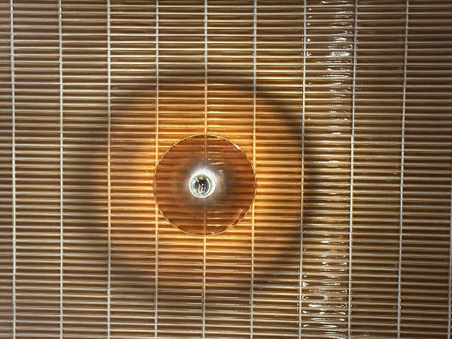 A flat-lay presents a rectangular tile pattern dominated by light brown hues. Individual tiles, arranged horizontally, form rows separated by thin, light-colored grout lines. A circular light fixture occupies the center. The fixture's metallic edges and bulb cast a diffused light that illuminates surrounding tiles, creating a radial gradient of brightness across the pattern. Some tiles exhibit glossy reflections, adding visual texture to the composition.