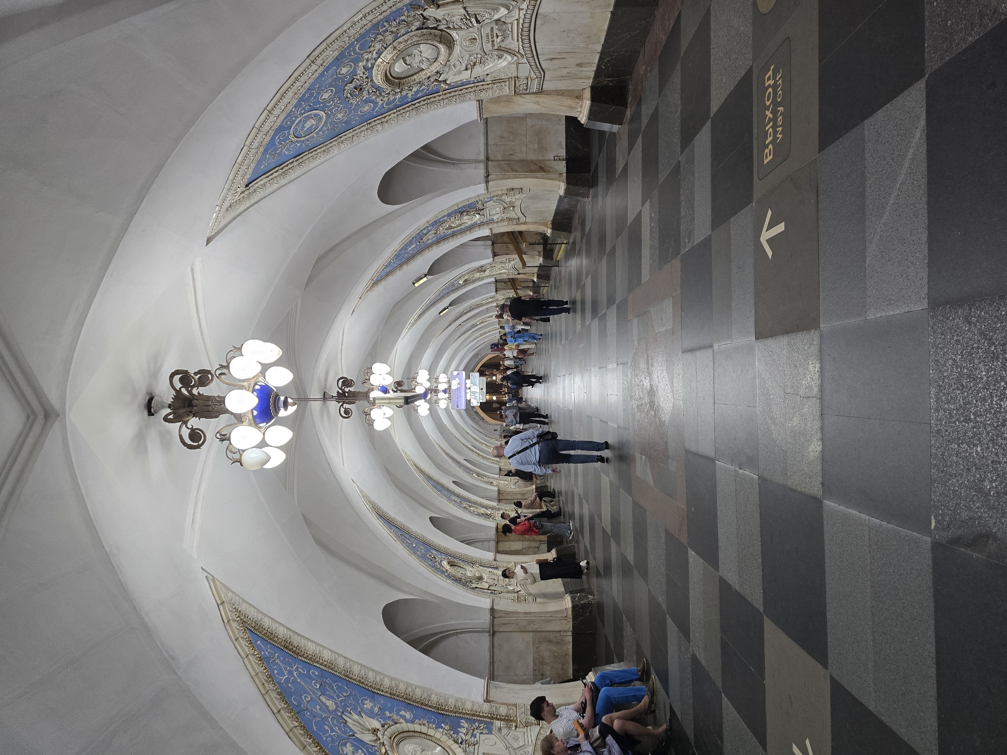 The image shows the Komsomolskaya metro station in Moscow, Russia. The scene is a long, arched hall with a checkered floor and ornate, white and blue ceiling decorations. Along the sides of the hall are benches where people are sitting and standing. Some people are looking at their phones, while others are talking or simply waiting. A sign on the floor reads "Выход / Way out" with an arrow pointing in one direction. Chandeliers hang from the ceiling. The perspective makes the hall appear to converge in the distance.