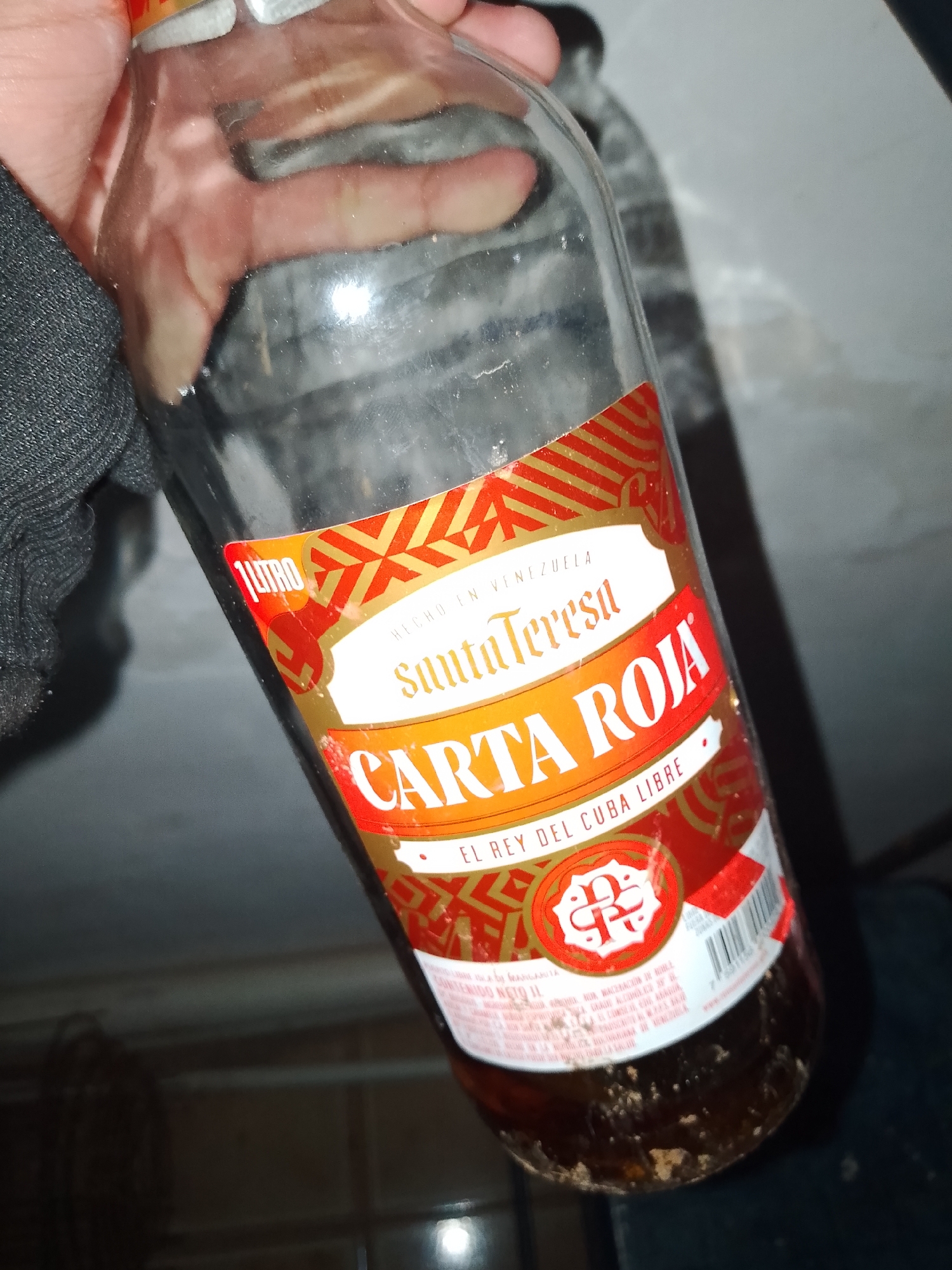 A clear glass 1-liter bottle of Santa Teresa Carta Roja rum, marked "HECHO EN VENEZUELA" and "EL REY DEL CUBA LIBRE," is held vertically by a human hand. The hand is partially covered by a dark, textured long-sleeved garment. The bottle contains a small quantity of dark brown liquid with visible particulate sediment at the bottom. The label is predominantly red and gold, featuring the brand "Santa Teresa" and product name "CARTA ROJA," along with a circular emblem and a visible barcode on the lower right side. The background consists of a light-colored, smooth surface in the upper right and a darker surface with faint linear patterns in the lower portion. The scene is illuminated, casting some reflections on the bottle. Location: Unknown, Unknown.