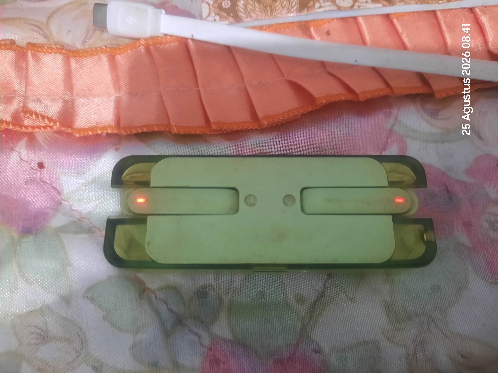 The image shows a close-up view of a light green electronic device, possibly a portable charger or power bank, lying on a patterned fabric surface. The device has two visible red indicator lights illuminated. To the right of the device is a white USB cable with a silver connector, and next to the cable is a bright orange ruffled fabric. The background fabric has a floral or abstract pattern with pink and white colors. A timestamp "25 Agustus 2026 08.41" is visible in the lower right corner of the image, indicating it was taken on August 25, 2026, at 8:41 AM. The scene is indoors, with soft lighting. There are no people or animals visible in the image. The location of Sukokerto, Indonesia, is not directly discernible from the visual elements of the image itself.