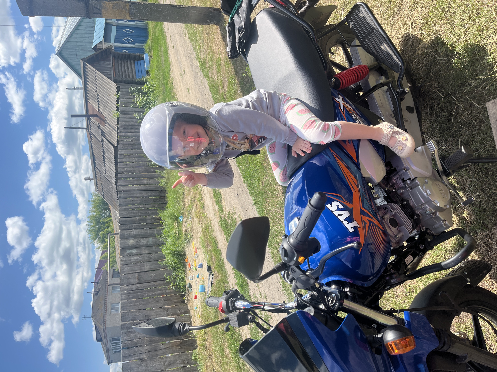 A young child wearing a silver helmet with a clear visor is sitting on the seat of a blue motorcycle. The child is dressed in a gray long-sleeved shirt with a graphic print and white pants with a watermelon pattern. They are smiling and pointing with their left index finger. The motorcycle is a TVS brand, with "TVS" visible on the blue fuel tank. The scene is outdoors, with a dirt path, dry grass, and a wooden fence in the background. Behind the fence, there are several rural-style wooden houses with peaked roofs and windows. The sky is blue with fluffy white clouds. The image appears to be taken on a sunny day. The overall impression is one of playful curiosity and a rural summer setting in Сухринское, Russia.