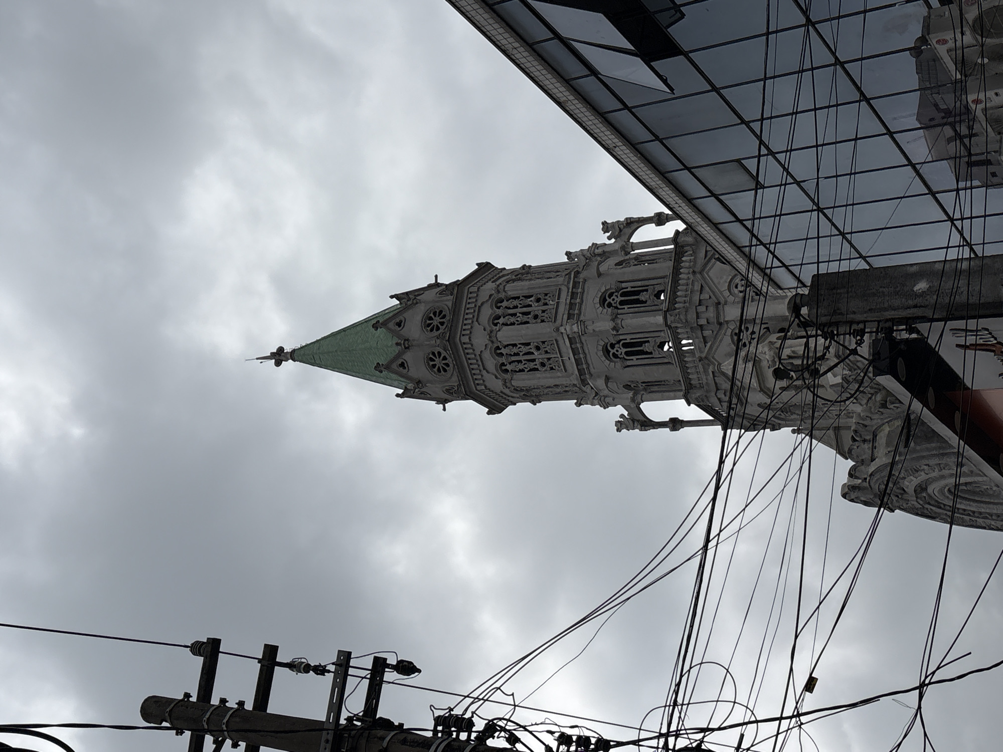The image shows the intersection of the Basilica of the Sacred Heart of Jesus and a modern building, framed by a telephone pole and a dense network of electrical wires. The basilica, a prominent landmark in the Bom Retiro neighborhood of São Paulo, Brazil, stands tall with its Gothic-style architecture, featuring intricate stone carvings and a distinctive green spire. Its ornate facade is partially obscured by the contemporary glass-fronted building, which reflects the cloudy sky and the surrounding urban landscape. The telephone pole, laden with wires, adds a sense of urban chaos and contrasts with the historical grandeur of the basilica. The overall scene captures the juxtaposition of old and new, religious and secular, in the vibrant cityscape of São Paulo. There are no people visible in the image.