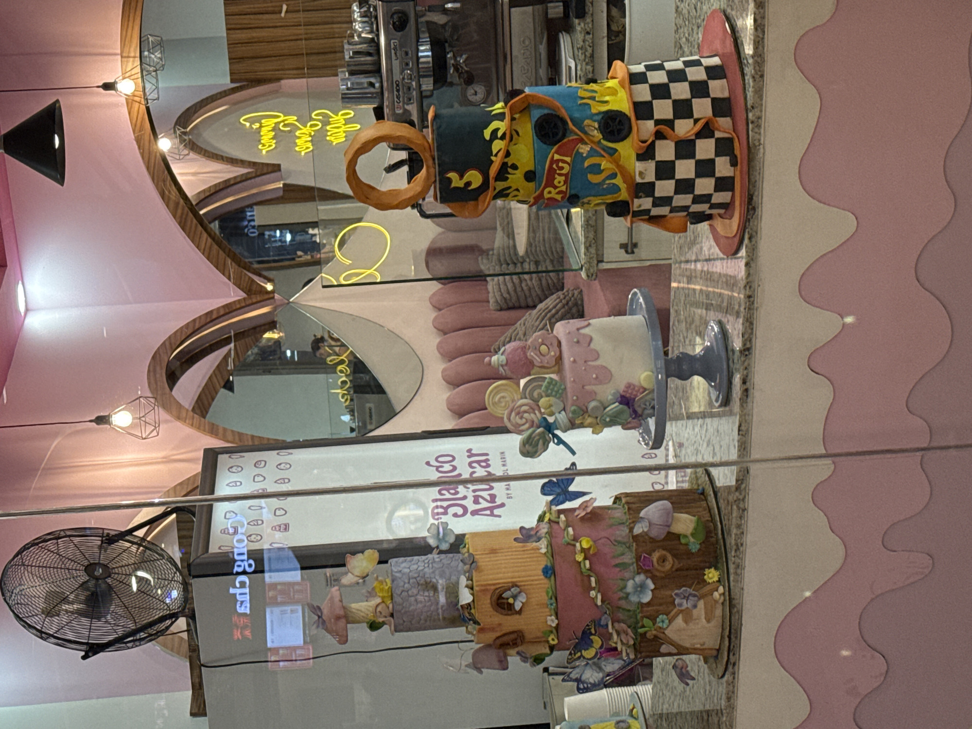 The image shows a display case of cakes at the "Blanco Azúcar" by Maol Marin bakery in Puebla, Mexico. There are three tiers of cakes in the case. The top cake is a multi-tiered design with racing-themed elements, including a donut-shaped piece, flames, and a checkered pattern. The middle cake is lighter in color, decorated with pastel candies and lollipops on a cake stand. The bottom cake has a woodland theme with butterflies, flowers, and mushrooms. The display case has a sticker with the bakery's name. A metal fan is visible in the corner of the image. The background of the image includes a coffee machine, neon signs, a reflective surface with pink walls.