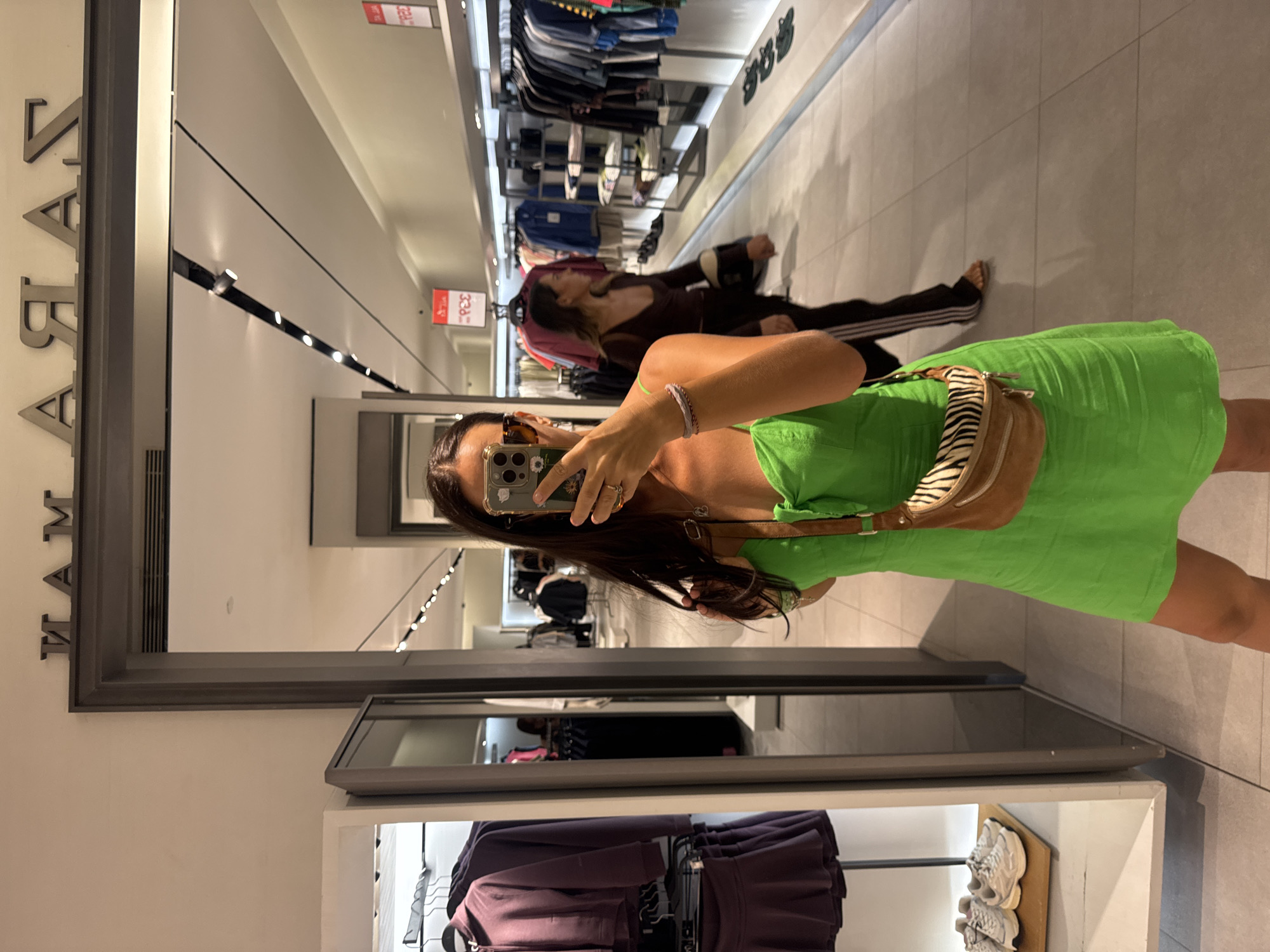The image depicts a woman taking a selfie in a Zara store in Kuta, Indonesia. She is wearing a bright green dress and a tan crossbody bag with a zebra print front. In the background, racks of clothing and mannequins are visible, along with other shoppers browsing the merchandise. The store appears to be well-lit and organized, with a modern retail aesthetic. The sign above the entrance reads "ZARA" in large, metallic letters. The woman is standing in front of a mirror, capturing her reflection as she takes the photograph with her smartphone.