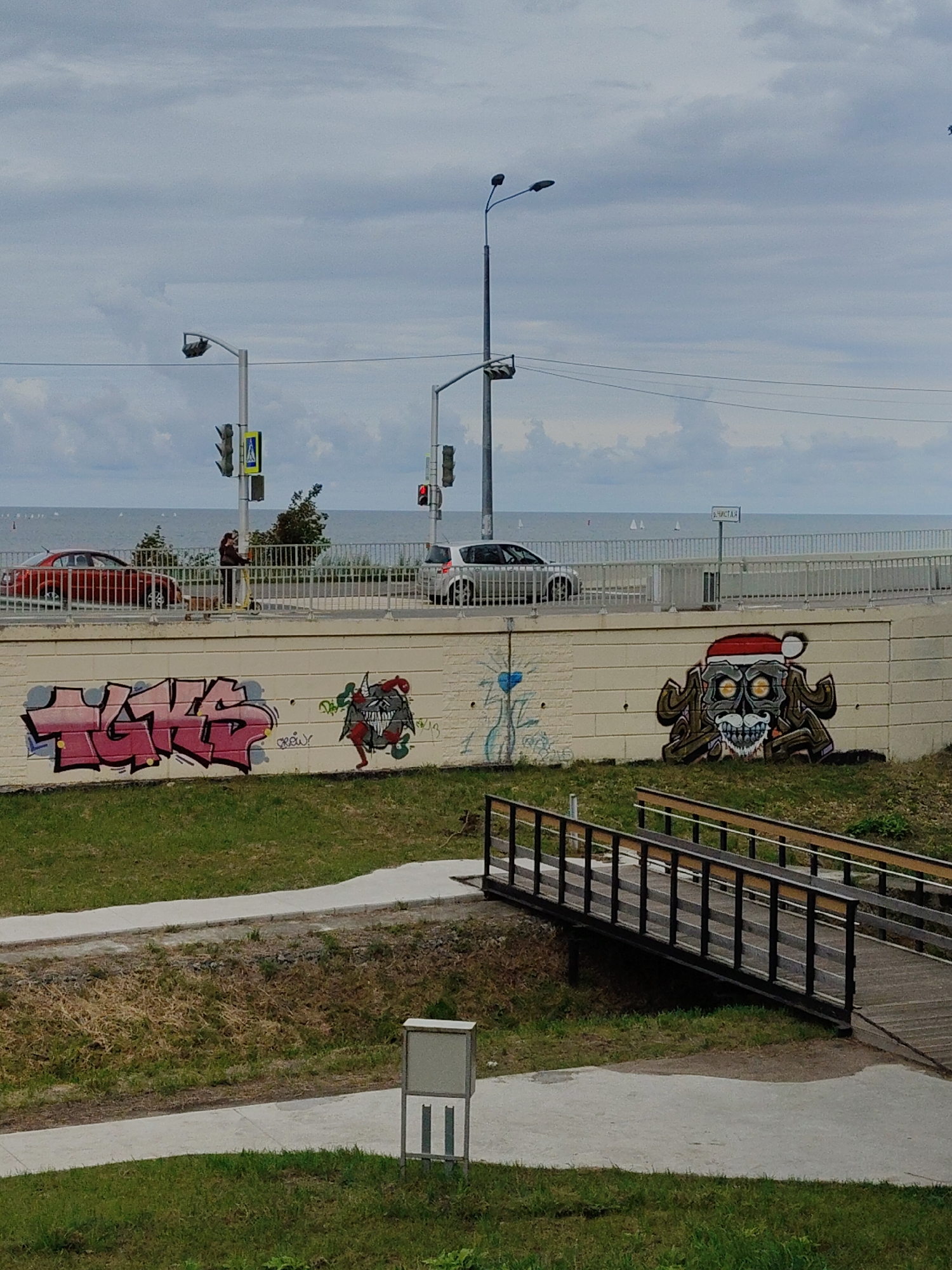 The image depicts a concrete retaining wall adorned with graffiti, situated near a body of water, likely the Baltic Sea, given the location of Pionersky, Russia. The graffiti on the wall includes a stylized pink word that appears to be "HATE" in large block letters, and to its right, a monstrous figure wearing a Santa hat, with a skull-like face, mustache, and dark, claw-like limbs. Behind the wall, a paved walkway runs parallel to a road where a red car and a white SUV are visible. Further back, a body of water stretches to the horizon under a cloudy sky. A pedestrian bridge with wooden planks and metal railings spans a grassy area in the foreground, leading away from the retaining wall and towards a concrete path and a grey information stand. The overall scene suggests an urban or semi-urban environment with elements of public space and artistic expression.