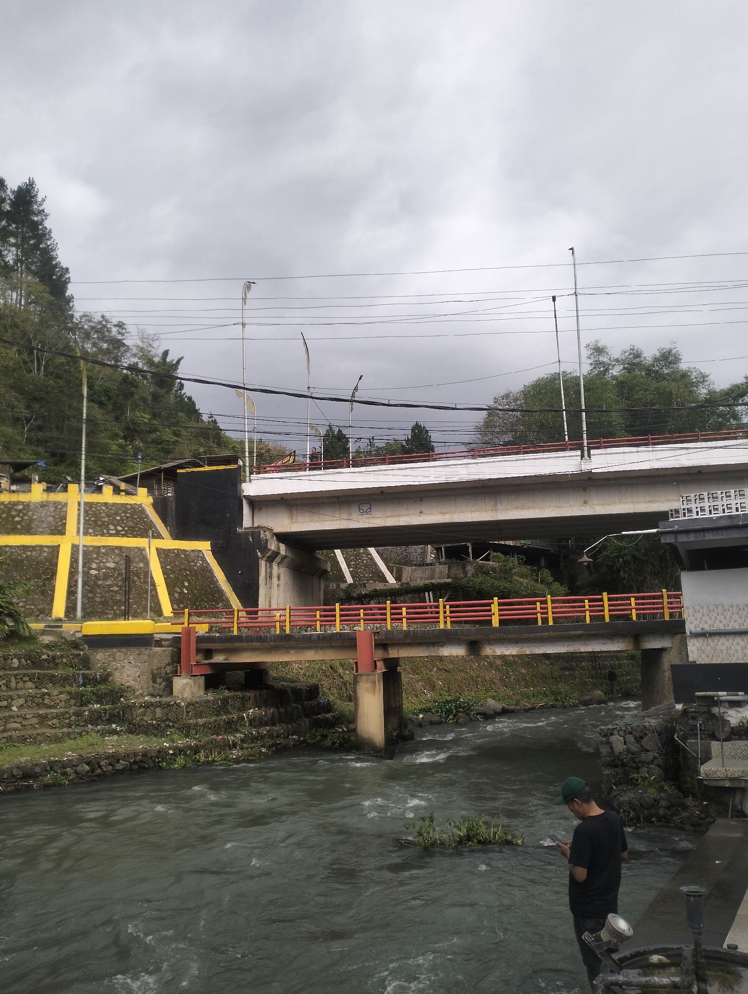 A man in a black t-shirt and green baseball cap stands by a river, looking down at his phone. He is on a concrete walkway that borders the dark, flowing water. In the background, two bridges cross the river. The lower bridge is a pedestrian bridge with red and yellow railings, while the upper bridge is a larger concrete structure with a white railing. A hillside covered in trees rises behind the bridges, with a retaining wall decorated with yellow and black patterns on the left side. The sky is overcast and gray, suggesting it may be a cloudy or rainy day. The scene appears to be in a natural or semi-urban setting with lush greenery. There is no visible text in the image.