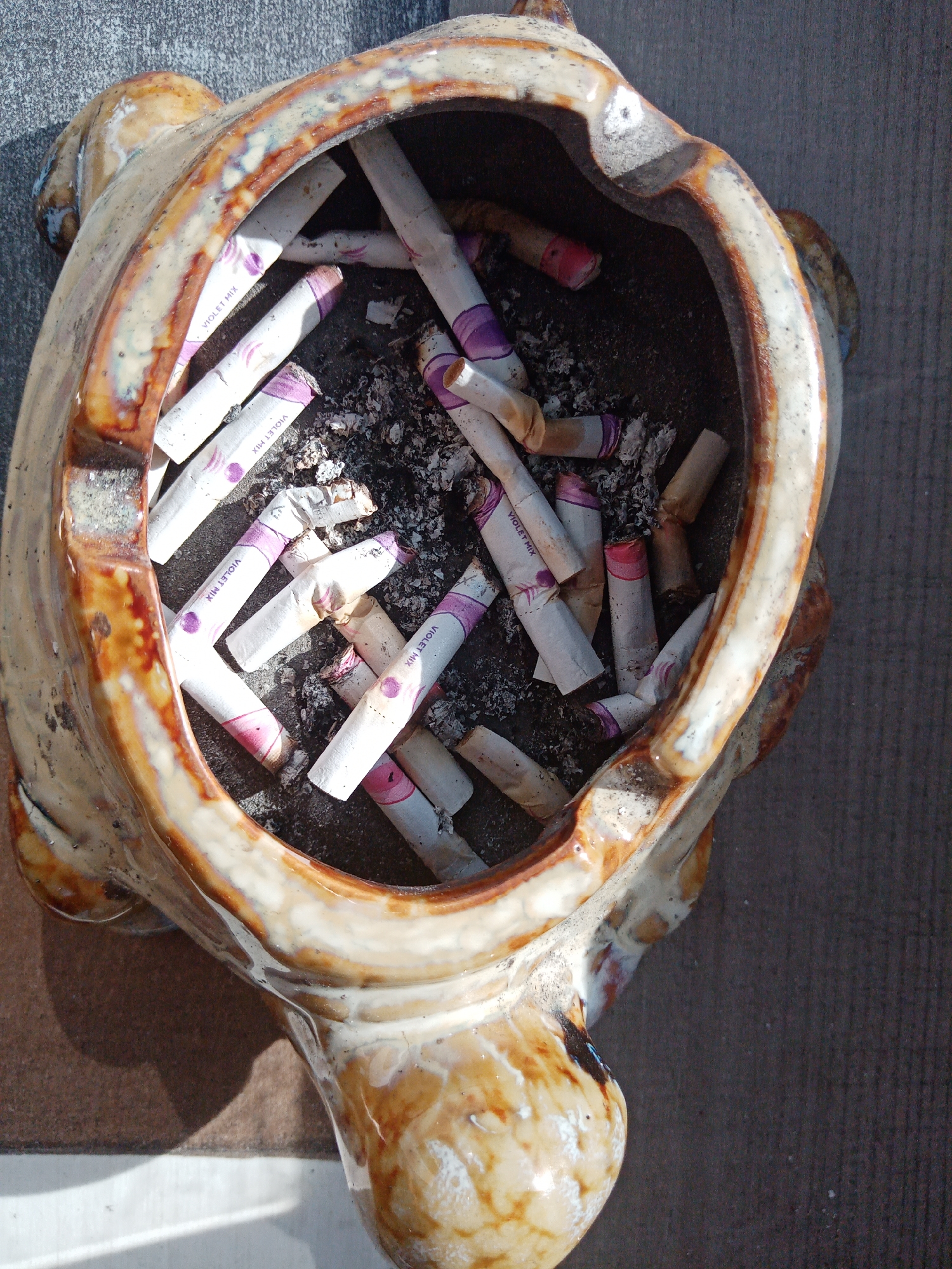 The image shows a tan and brown ceramic ashtray, shaped roughly like a turtle, filled with cigarette butts. The cigarettes appear to be of the "Violet Mix" brand. The ashtray is sitting on a gray surface. No people are visible. The location is Elektrostal, Russia.