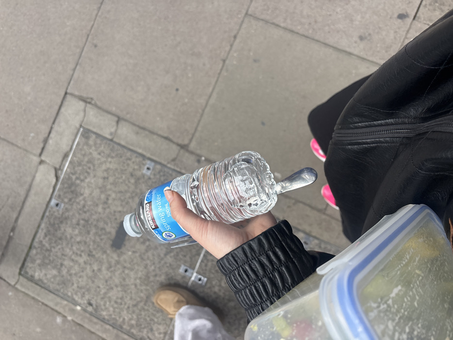 The scene depicts a first-person, downward-facing perspective of a pedestrian area in the city of London, United Kingdom. In the foreground, a person's left hand, wearing a black, textured long-sleeved garment or jacket, holds a clear plastic 500ml Kirkland brand spring water bottle. The bottle is mostly full, and its base shows a distinct molded design or reflection resembling a large water drop. To the immediate left, a portion of a transparent plastic food container with a blue-rimmed lid is visible, held or positioned near the person's left hip area, suggesting it contains food. The person's lower body is also visible, wearing dark trousers or a skirt and pink and white athletic shoes. In the upper left background, the right foot of a second individual is visible, wearing a tan loafer-style shoe and white hosiery. The ground is a grey paved surface, constructed of multiple concrete or stone slabs with visible seams and cracks. A square, dark grey metal utility access cover, secured by four bolts, is embedded in the pavement in the upper left quadrant of the image. The lighting appears overcast, creating soft, diffused illumination without strong shadows.