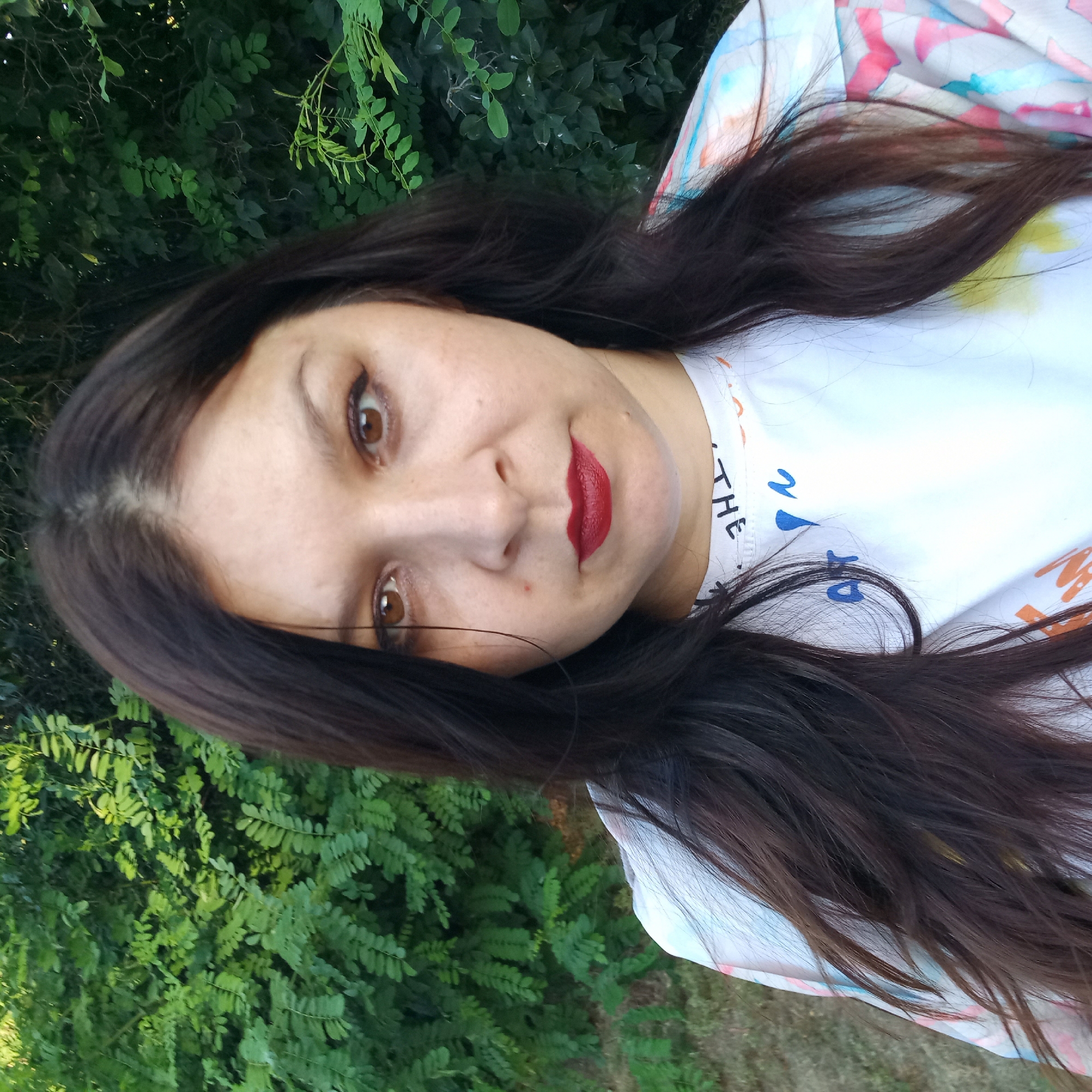 A woman with dark hair and brown eyes, wearing a white t-shirt with a colorful abstract pattern and text, poses for a selfie outdoors. She is positioned in the foreground, with lush green foliage forming the background. The location is not specified as Klyntsi, Ukraine.