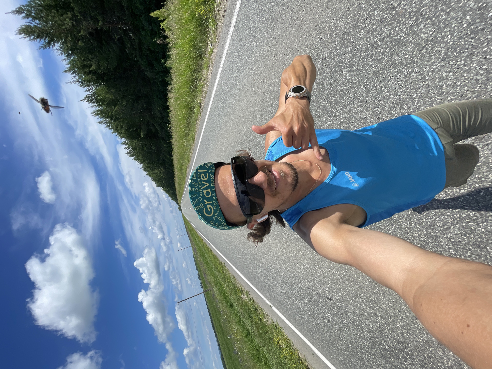 An outdoor, eye-level, medium-shot selfie captures a smiling man on a paved road bordered by green fields and a line of pine trees under a bright blue sky with scattered white clouds. The man, who appears to be in his late 30s or early 40s, has long, brown hair pulled back and wears large, dark sunglasses, a blue tank top with a "Jako" logo, khaki pants, and a green baseball cap with the word "Gravel" on it. He has white wireless earbuds in his ears and is making a "shaka" hand gesture with his left hand and a thumbs-up with his right hand. A silver smartwatch is visible on his right wrist. In the upper right quadrant of the image, a large insect, possibly a dragonfly, is captured mid-flight. The road stretches into the distance, with power lines visible on the left side.