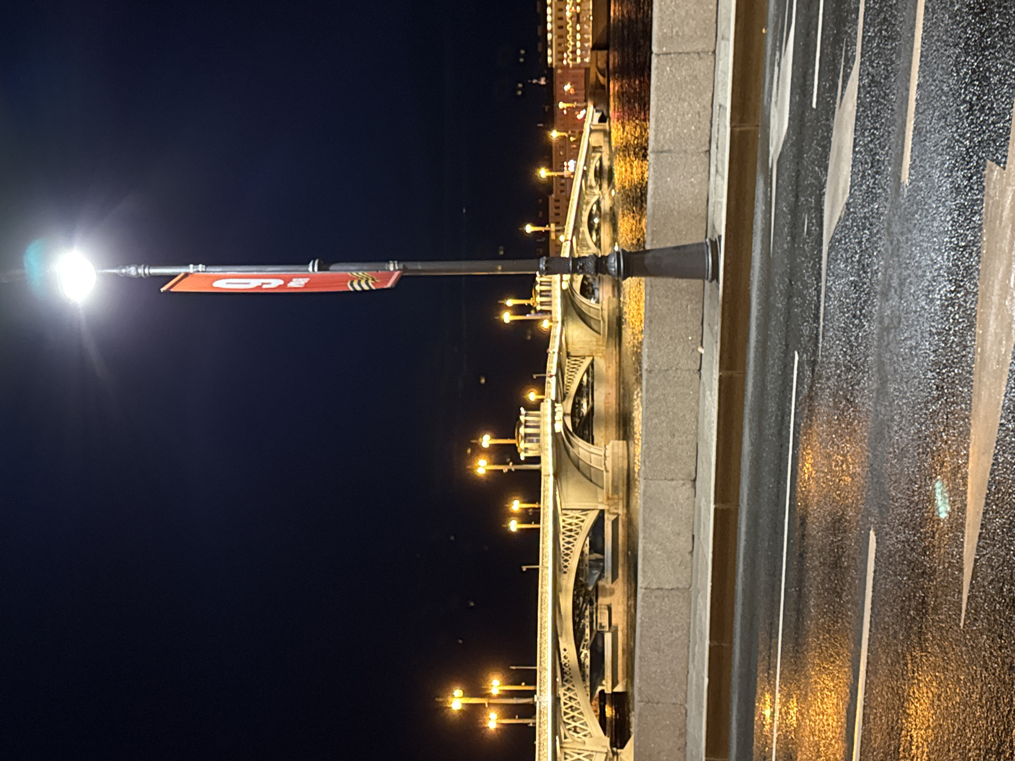 The image captures a nighttime view of a bridge in Saint Petersburg, Russia. The perspective is from the side of a road with lane markings. A black lamppost with a bright white light and a red banner displaying '9 мая' is visible. The bridge itself is adorned with rows of lights reflecting on the water below. Buildings are discernible in the background, also illuminated at night. The road surface is wet, reflecting the surrounding light.