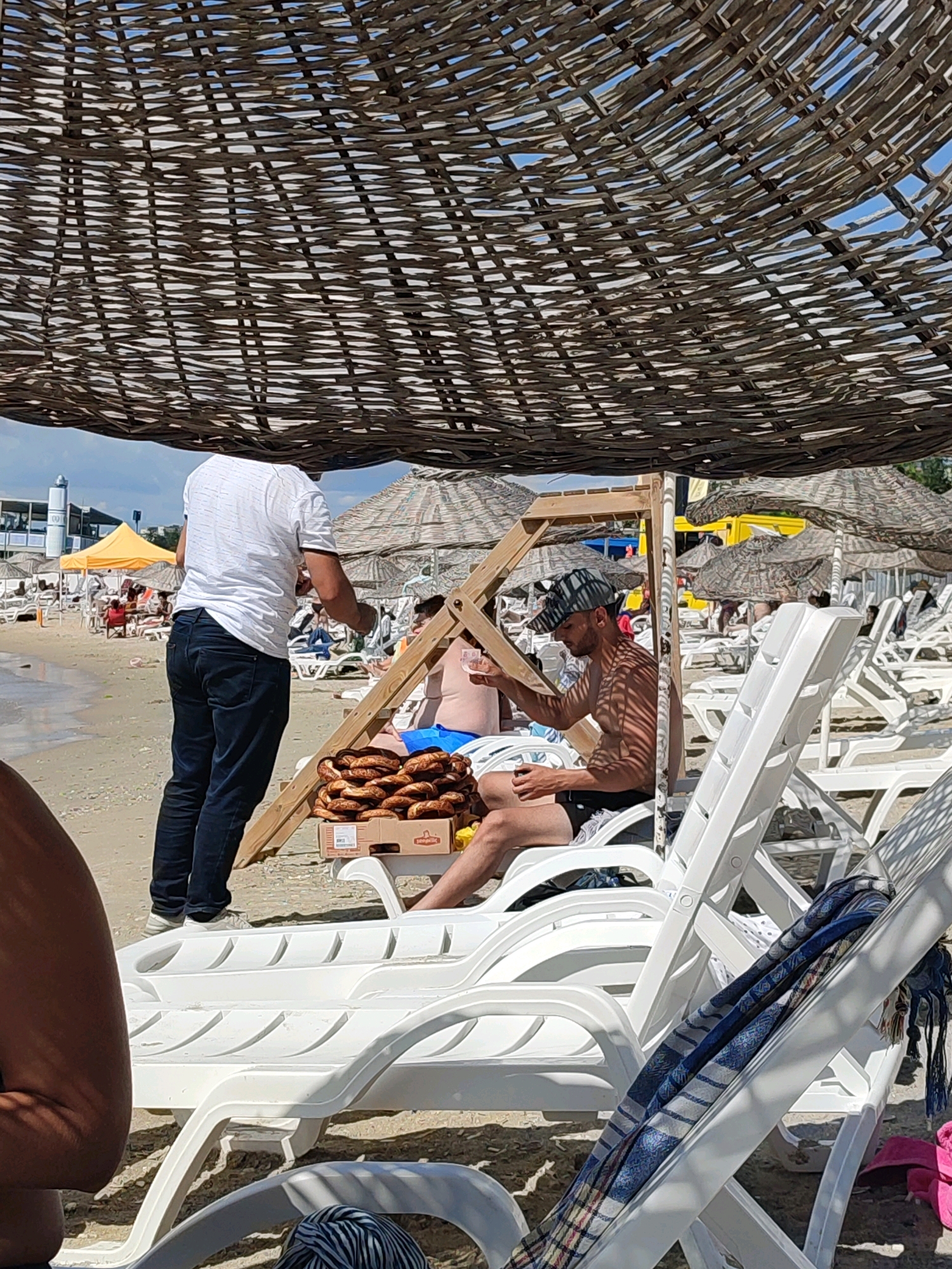 The image depicts a beach scene, likely in Kadıköy, Turkey, under a woven straw canopy. A man in a white t-shirt and dark jeans stands, facing away from the camera, holding a transparent cup. In front of him, on a wooden stand and a white plastic lounge chair, is a large cardboard box filled with numerous simit (Turkish bagels). To the right, a shirtless man wearing a patterned cap and black shorts sits on a white lounge chair, also holding a transparent cup. He is partially shaded by the canopy, and sunlight casts a striped shadow pattern on him. In the background, the beach is lined with white lounge chairs and umbrellas, with people scattered across them. A yellow tent and a light-colored building are visible in the distance. The sandy beach meets the water under a bright blue sky.